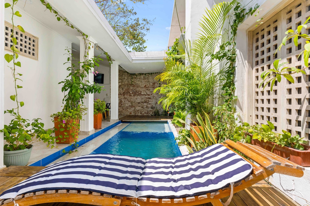A private pool is framed by lush greenery and potted plants, creating a serene outdoor space. A lounge chair positioned beside the pool features a striped cushion, inviting guests to relax. Natural light brightens the area, enhancing the tranquil atmosphere.