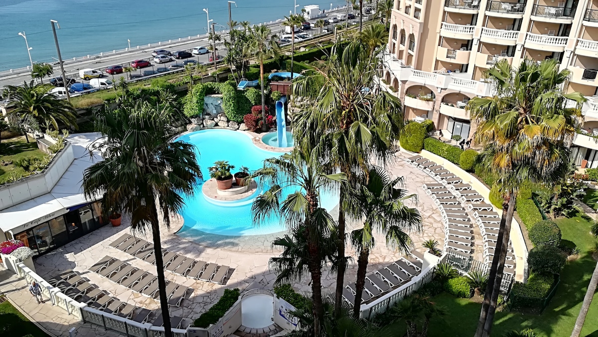 The image captures a vibrant pool area surrounded by lush palm trees. A large, inviting pool features a fountain, with numerous sun loungers arranged around it. The ocean can be seen in the background, creating a serene backdrop to the landscaped gardens.
