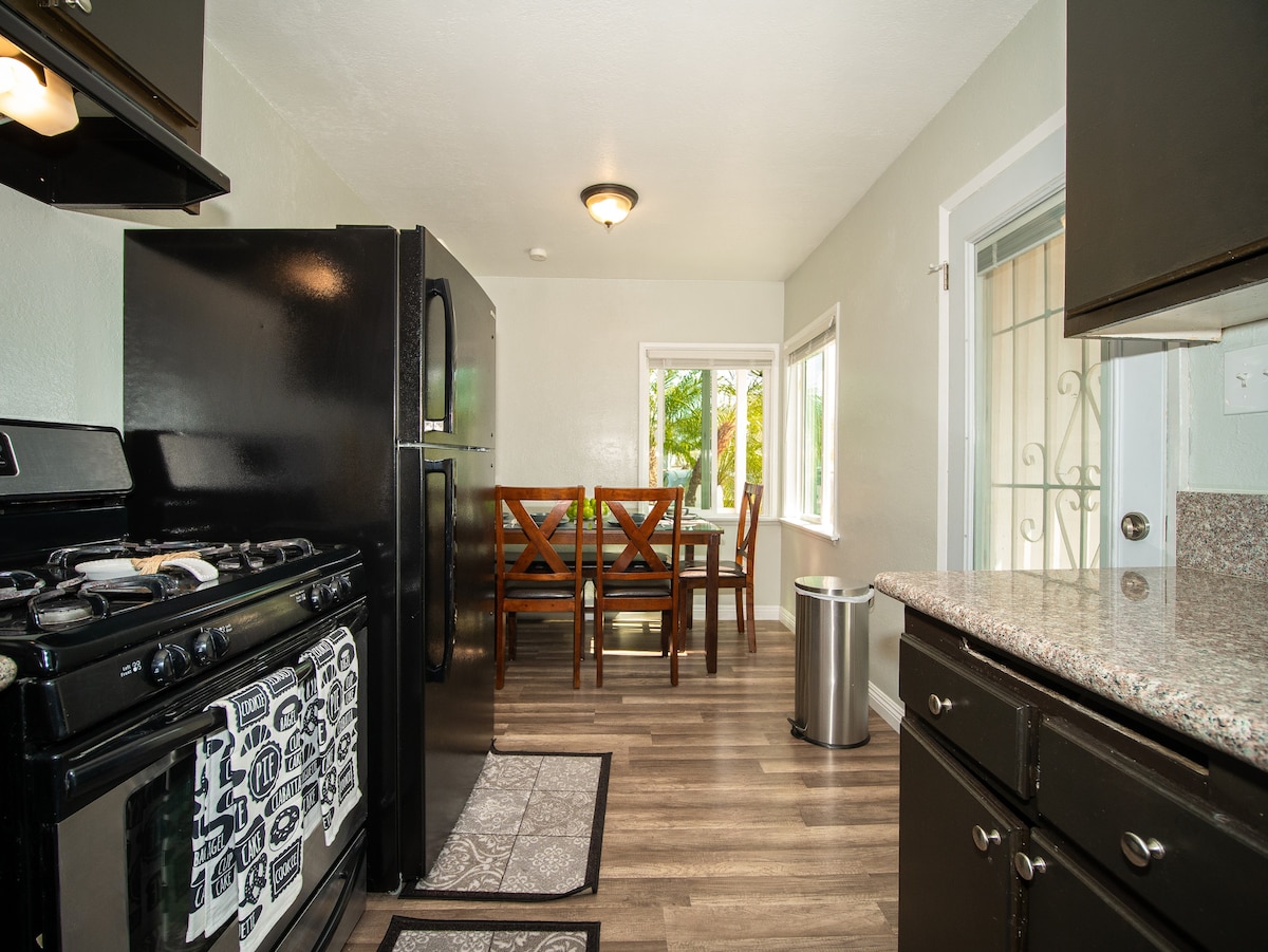 The kitchen features black appliances, including a gas stove and refrigerator. A granite countertop is visible, alongside a wooden dining table with four chairs. Natural light enters through a nearby window, illuminating the space.