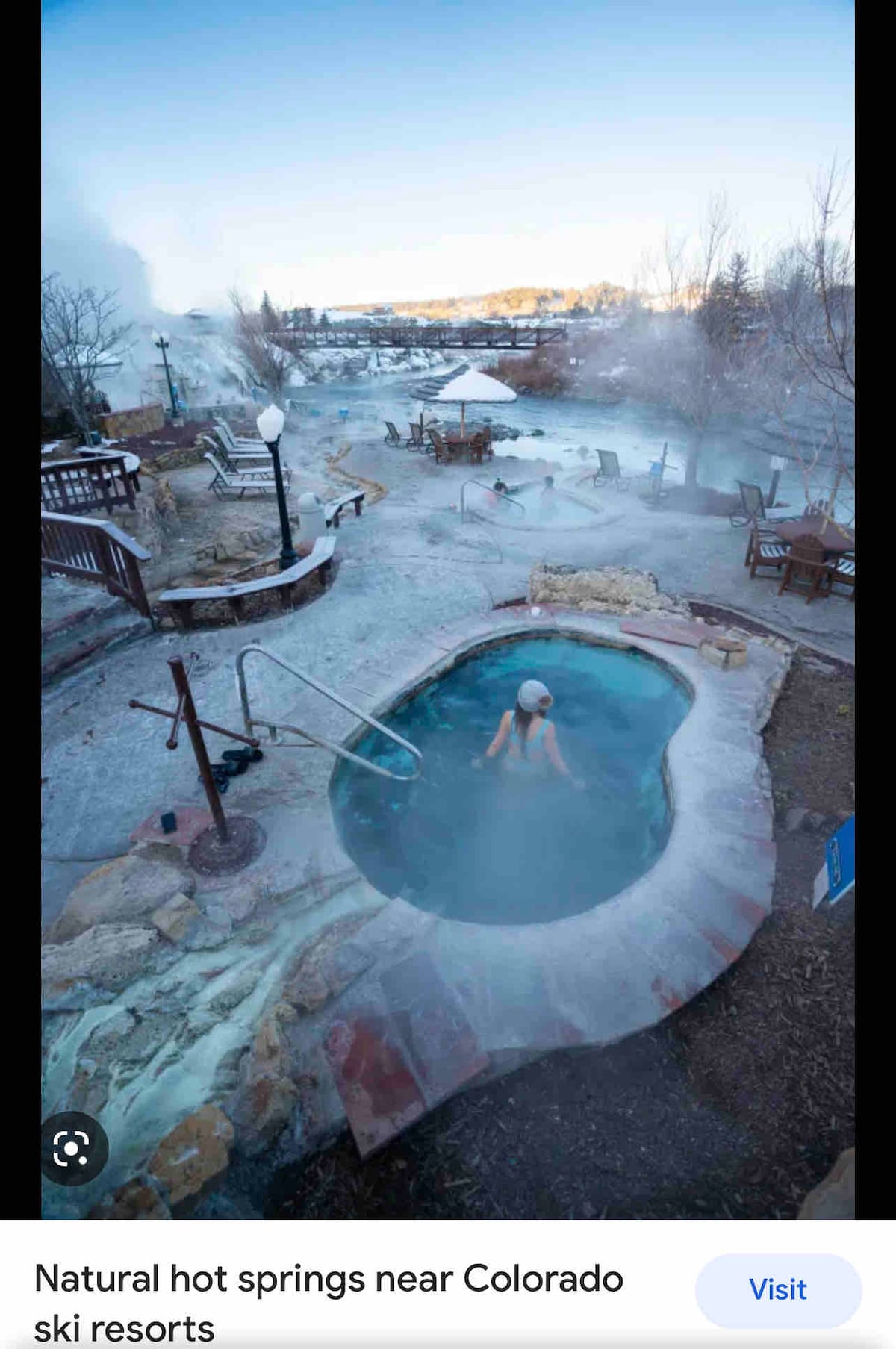 A natural hot spring is depicted, featuring steam rising gently in a snowy landscape. Surrounding wooden decks with seating are visible, along with frosted trees and distant mountains. A person is seen relaxing in the warm water, creating a serene atmosphere amidst the winter scenery.