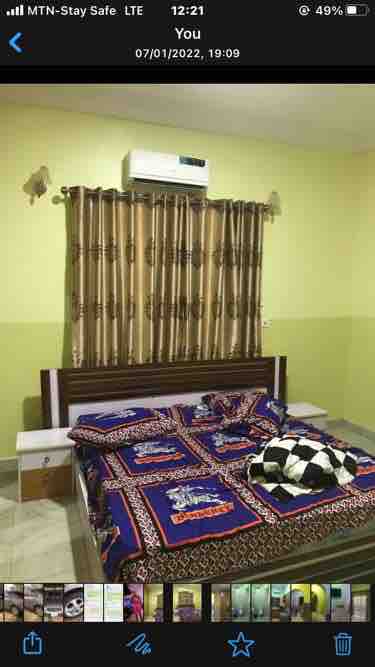 A cozy bedroom is presented with a spacious bed covered in a colorful patterned blanket. An air conditioning unit is mounted overhead, while light filters in through a window adorned with golden-brown curtains. A nightstand is positioned beside the bed.