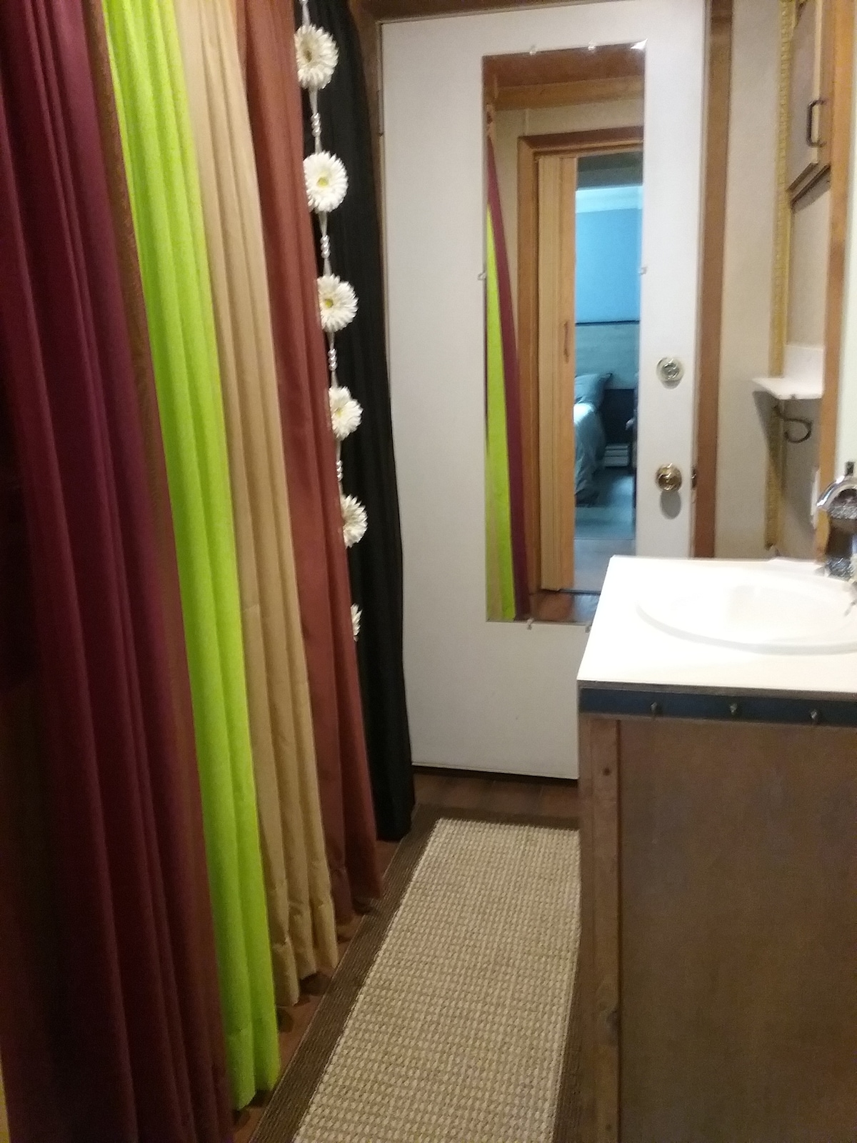 A compact bathroom area features a mirrored door that reflects the opposite bedroom. Vibrant curtain panels in shades of red, green, and black create a colorful division between spaces. A small sink is positioned against a wooden counter with a textured mat laid on the floor.