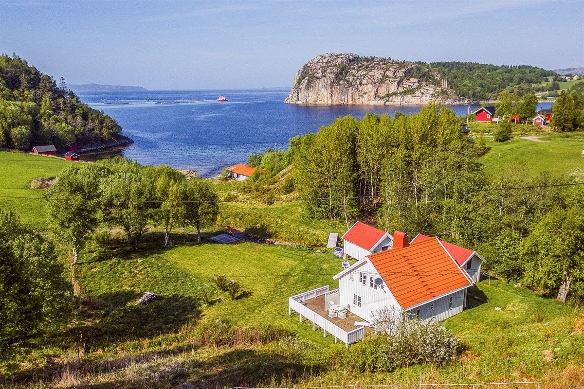 A panoramic view of a white house with a red roof is framed by a lush green landscape. The sea glistens in the background, along with a large rocky outcrop. Nearby, a few red buildings dot the scenery, emphasizing the serene rural setting.