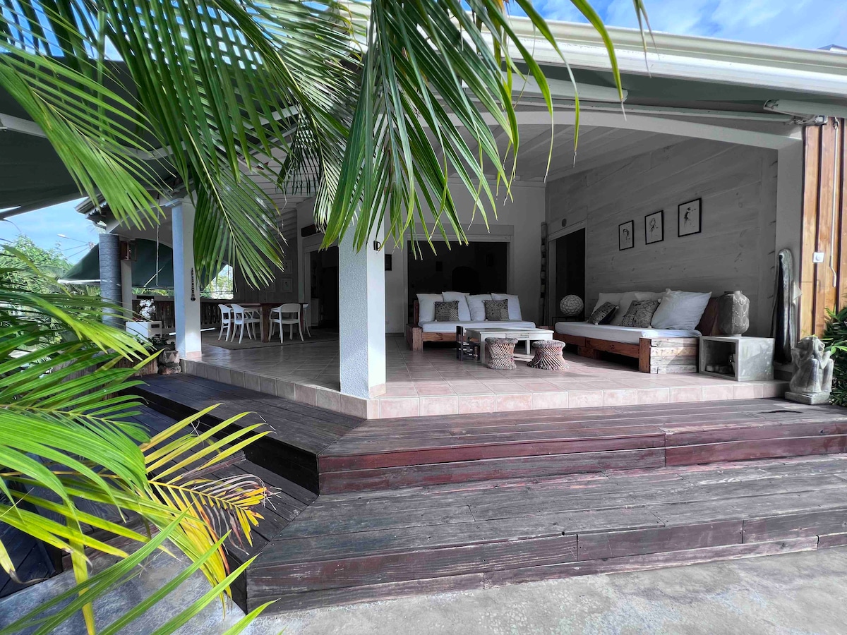 An outdoor seating area is visible under a covered terrace, featuring wooden furniture and cushions. Steps lead up to the space, surrounded by lush greenery. A spacious interior is seen through open doors, contributing to a relaxed ambiance.