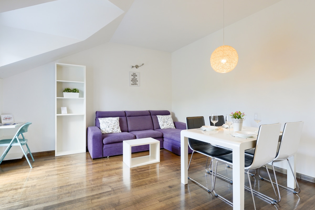 A cozy living area features a purple sofa paired with a white coffee table. A dining table with black chairs is set for meals, complemented by a simple pendant light. A shelf with decorative items enhances the space, while wooden floors reflect natural light.