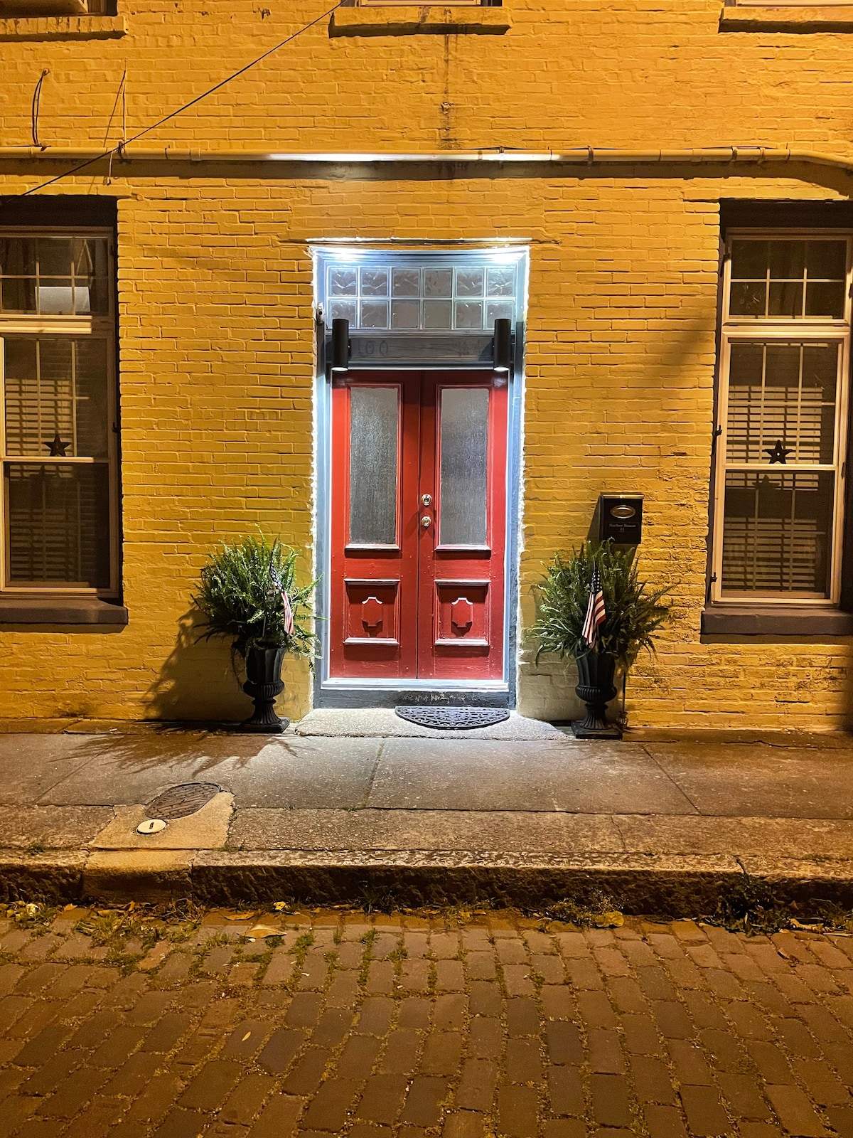 A striking red door is featured in the center of a brick facade, framed by large windows with shutters. Flanking the door are two potted plants, adding greenery to the entrance. The warm glow from inside casts an inviting light onto the cobblestone street.