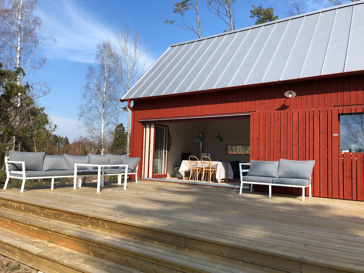 The exterior of a red barnhouse is displayed, featuring a spacious wooden deck. Comfortable seating arrangements, including two sofas, are positioned on the deck, with large glass doors leading to an indoor dining area visible. Natural surroundings with trees enhance the inviting outdoor space.