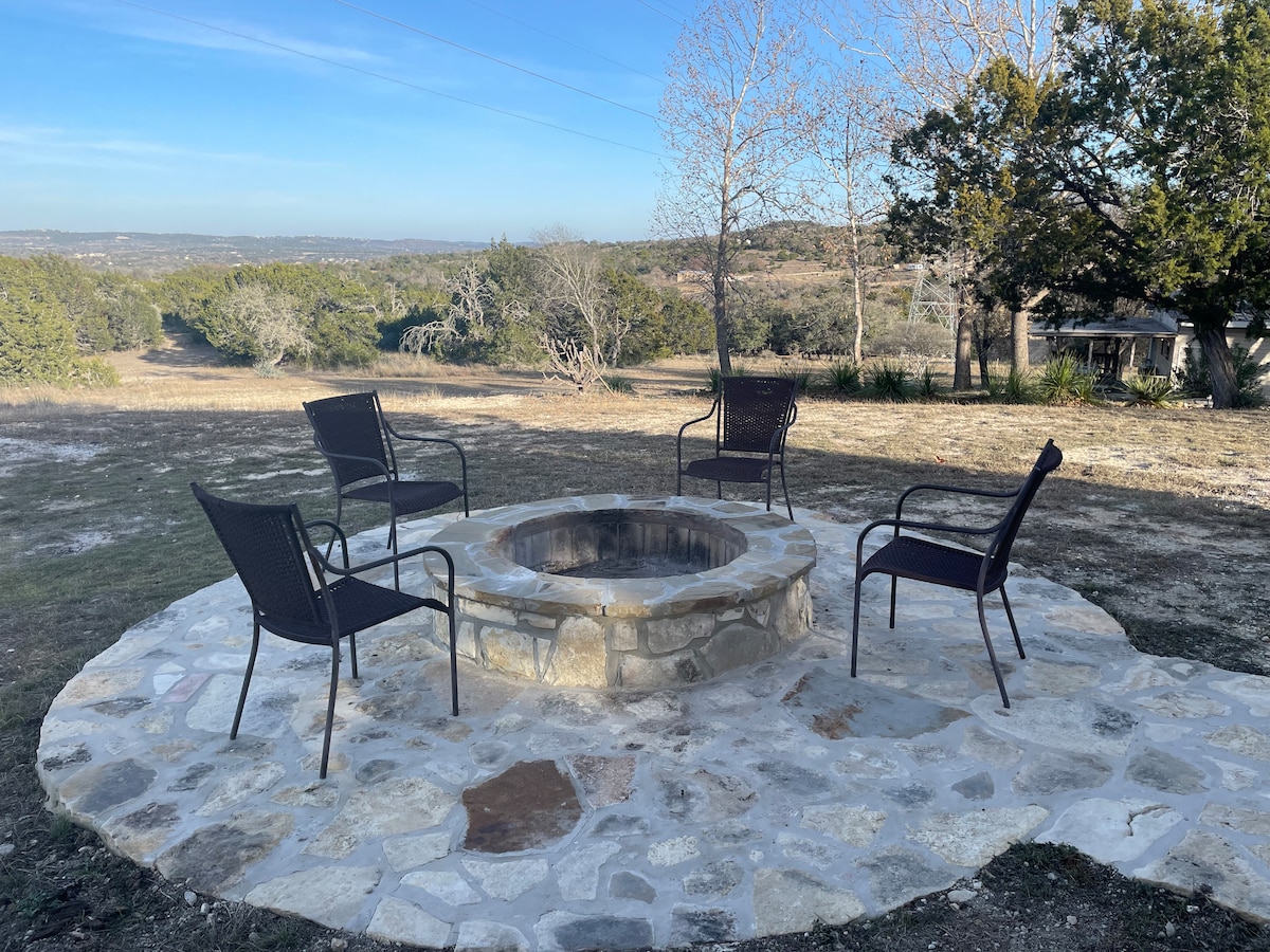 A circular stone firepit is surrounded by four black chairs situated on a stone patio. The expansive view of rolling hills and greenery is visible in the background, complemented by a clear sky. Trees dot the landscape, adding to the serene atmosphere.
