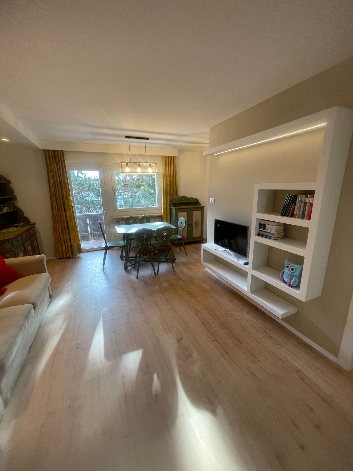 A bright dining area is highlighted by large windows allowing natural light to fill the space. A modern white bookshelf is mounted on the wall, complemented by a dining table and green chairs. A cozy sofa adds comfort to the inviting atmosphere.