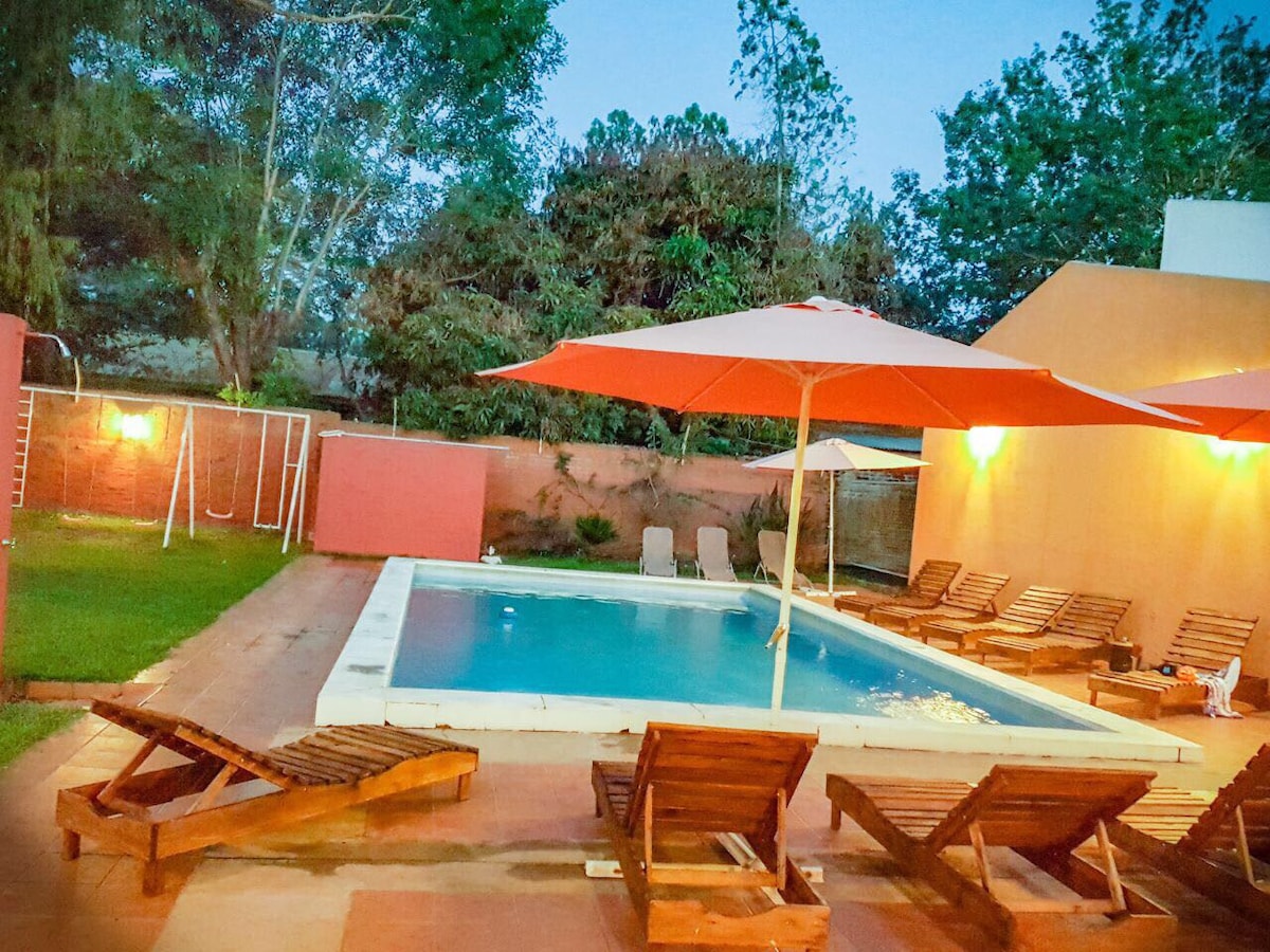 The outdoor pool area features a rectangular swimming pool surrounded by wooden lounge chairs. Large umbrellas provide shade, and a well-maintained lawn is nearby, with soft lighting enhancing the serene ambiance. A swing set is partially visible in the background.
