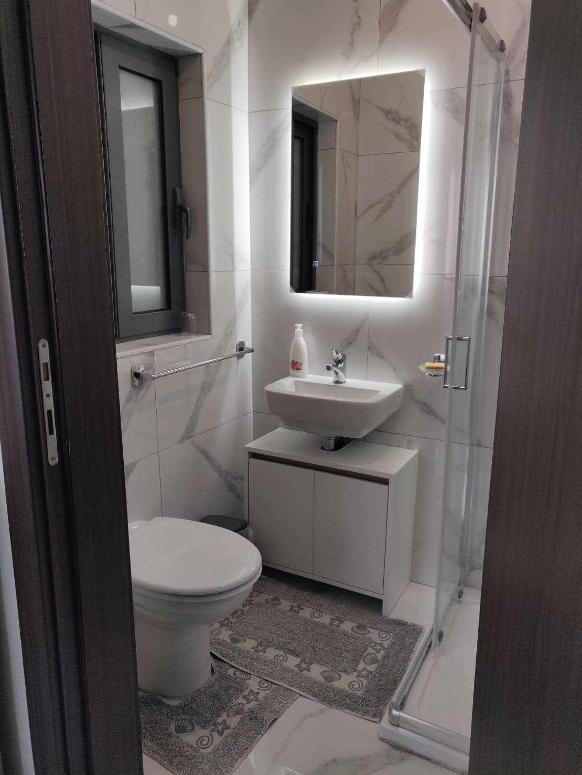 A compact bathroom features modern tiles with a minimalist aesthetic. A wall-mounted mirror and sink are complemented by a sleek shower area, while a small rug adds a touch of comfort. Natural light enters through a window, enhancing the clean design.