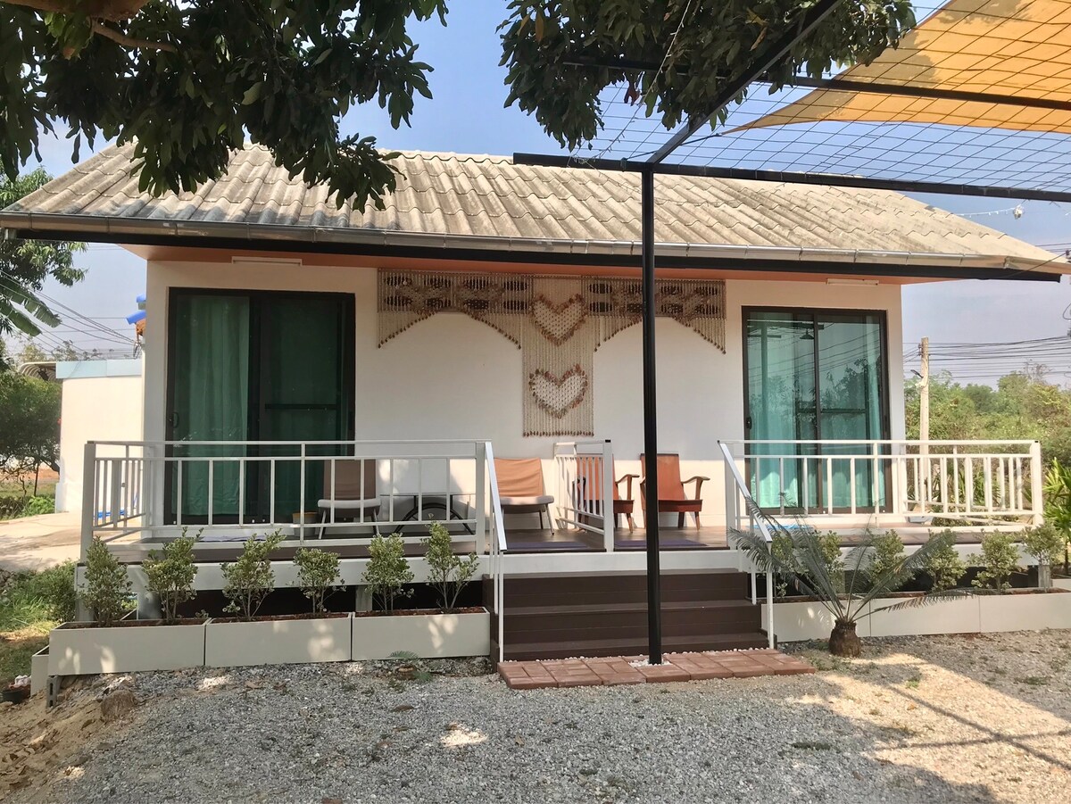 A quiet retreat is showcased, featuring a single-story structure with a light-colored exterior. Expansive glass doors open onto a welcoming porch with wooden seating and lounge chairs. Lush greenery borders the home, enhancing the serene atmosphere of the surrounding garden.
