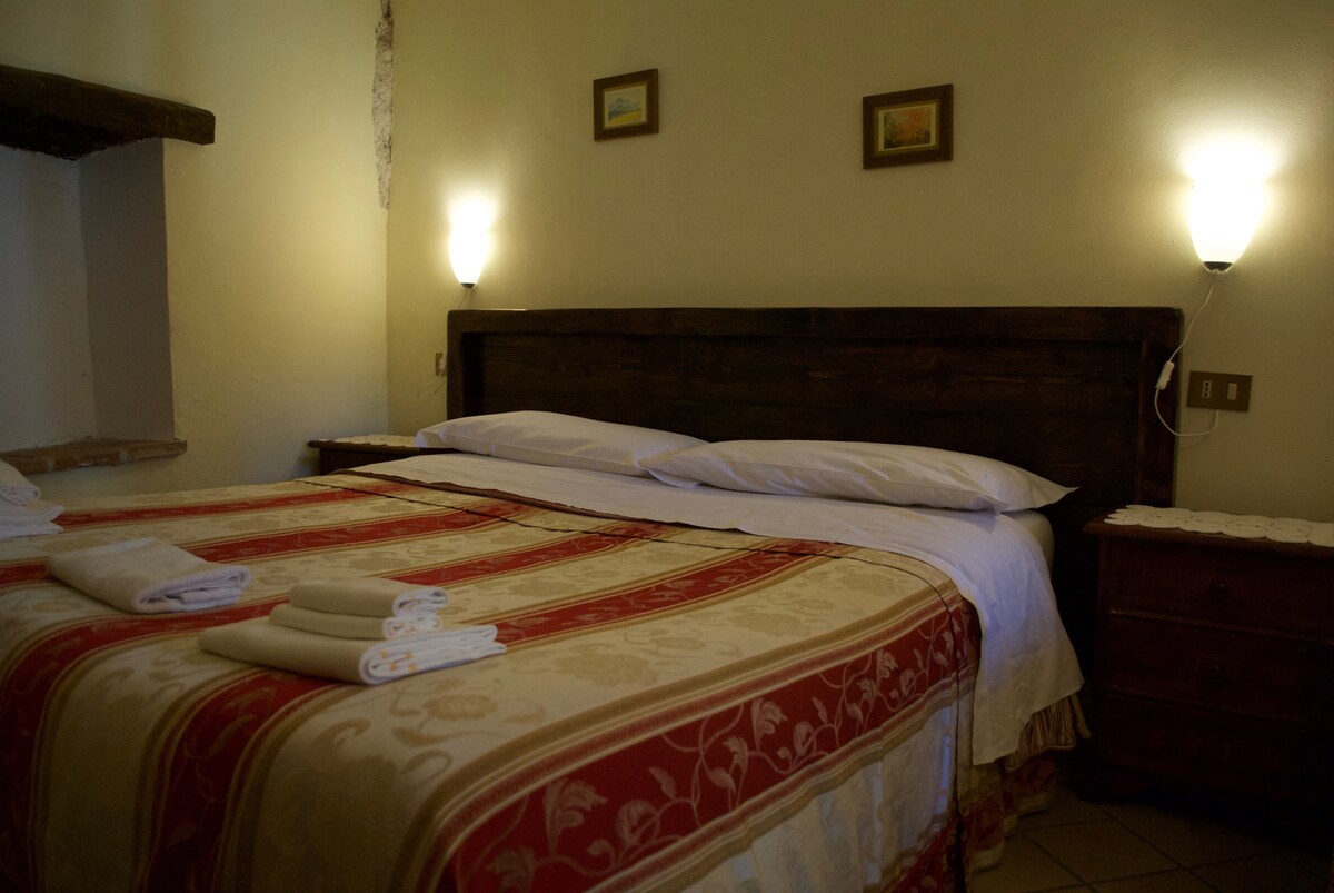 A cozy sleeping area features a spacious bed adorned with a patterned red and cream bedspread. Soft white linens are neatly arranged with towels placed at the foot of the bed. Subtle wall lights provide gentle illumination, creating a warm ambiance.