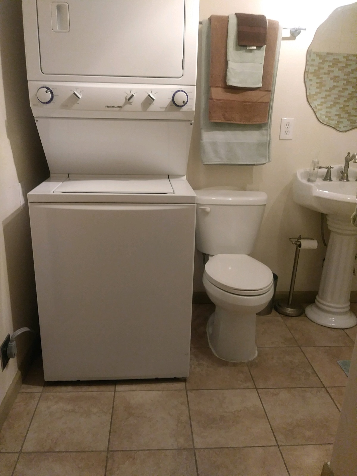 A compact bathroom is shown, featuring a stacked washer and dryer unit, neatly positioned beside a white toilet. A pedestal sink is equipped with a mirror above it, while towels are displayed on a rack nearby, adding to the organized arrangement of the space.