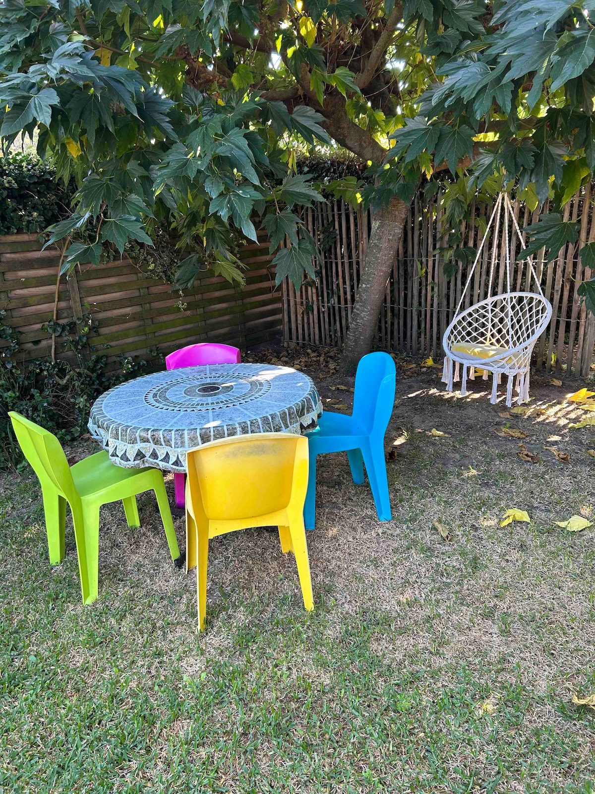 A colorful outdoor seating area features a round patterned table surrounded by chairs in vibrant hues of pink, blue, green, and yellow. A hanging swing chair is positioned nearby, sheltered by a leafy tree. The grass is well-maintained, providing a relaxing space.