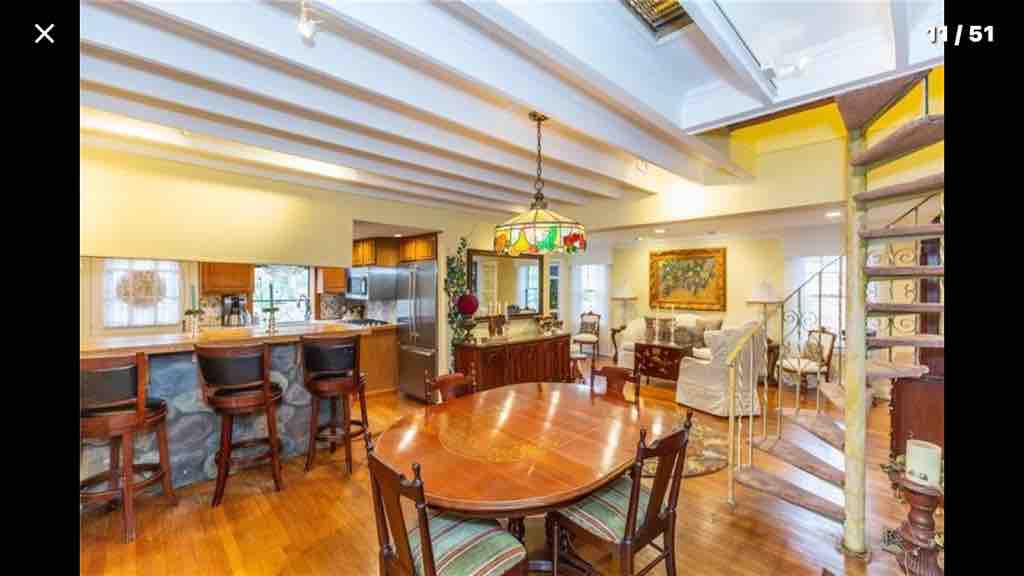 A spacious dining area features a round wooden table surrounded by six chairs. Bright light floods the room from large windows, highlighting the warm wooden floors. The kitchen area is visible in the background, equipped with modern appliances and a bar counter with stools.
