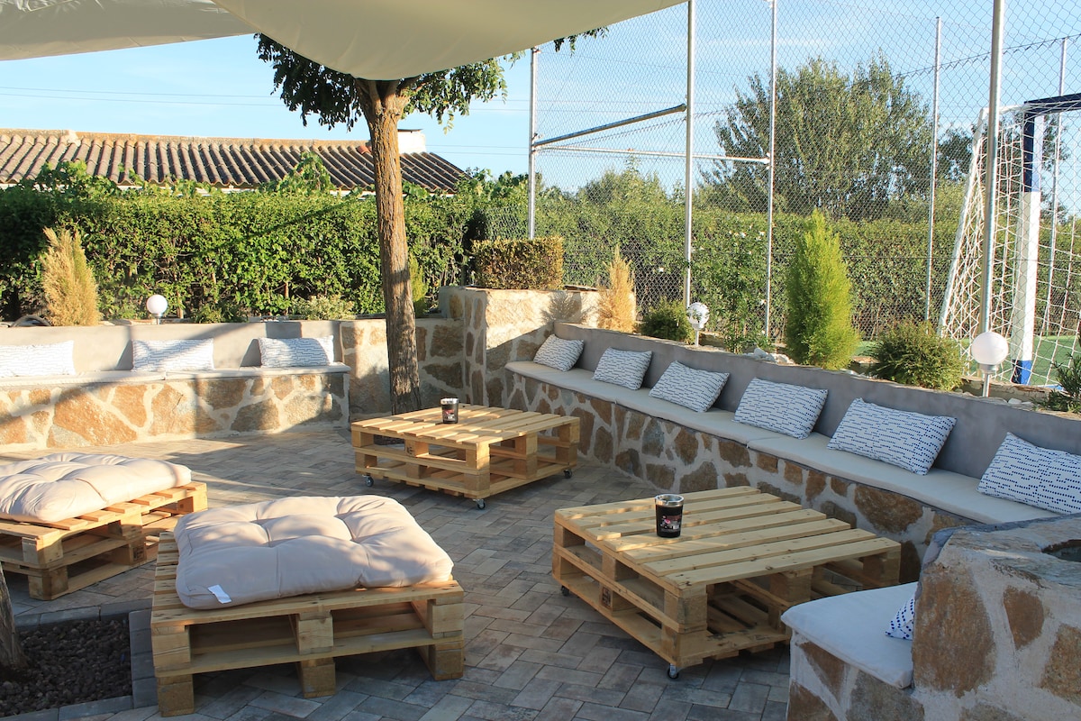 An outdoor seating area features cozy, low wooden pallet tables surrounded by cushioned benches. A shaded canopy offers protection from the sun, while lovely plants and greenery frame the space, creating a relaxed environment for gatherings.