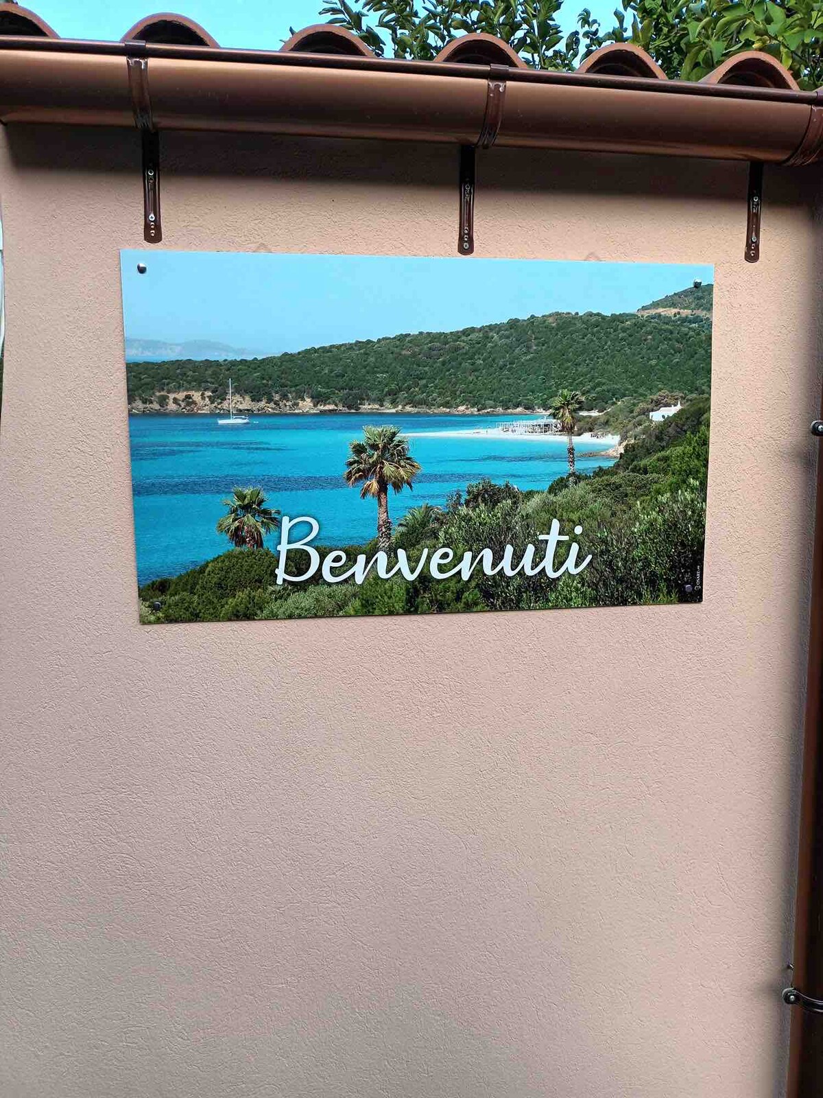 A welcoming sign hangs on a light-colored wall, featuring the word 'Benvenuti.' In the background, a serene coastal landscape with lush greenery and palm trees is depicted, complemented by clear blue waters and a tranquil sandy beach.