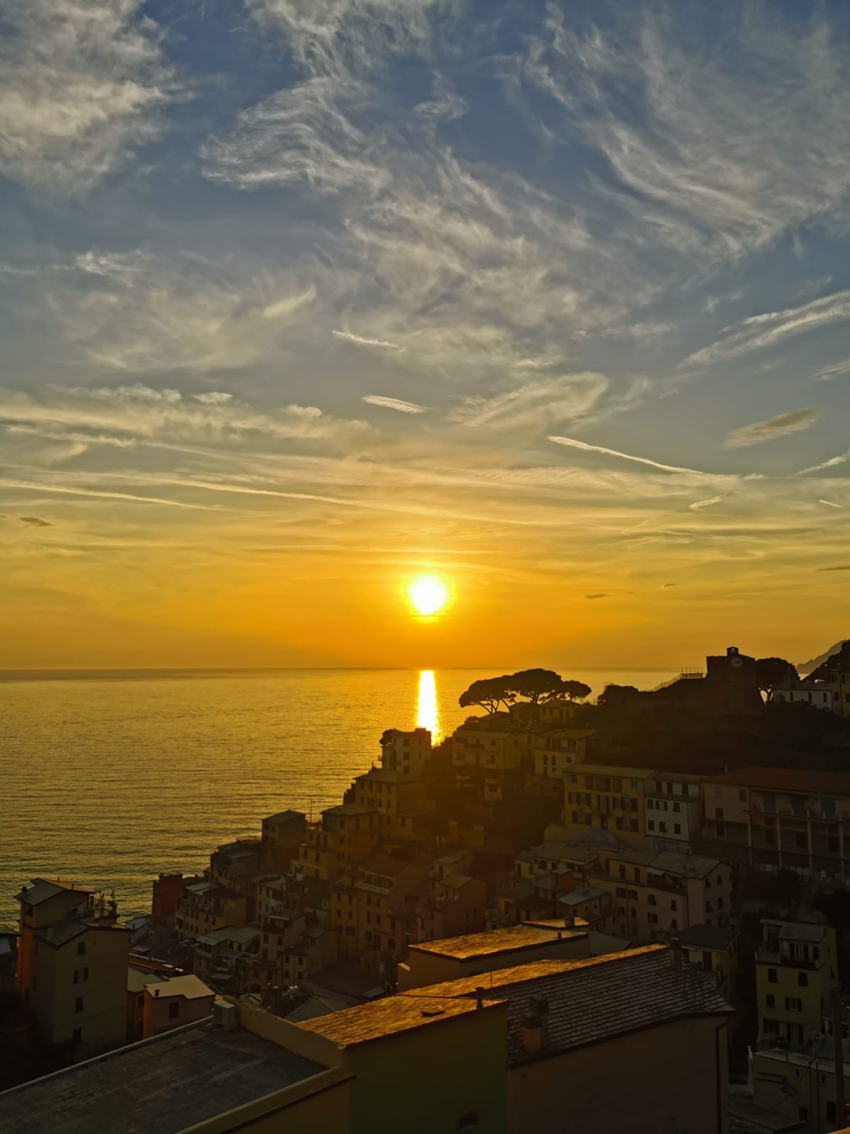 A stunning view captures the sun setting over the shimmering sea, casting golden reflections on the water. The silhouette of Riomaggiore’s colorful houses provides a contrast against the vibrant sky, highlighted with clouds and streaks of light.