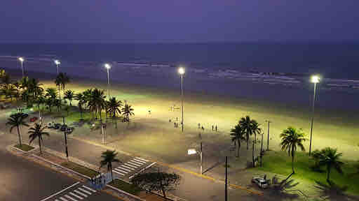 The image captures a serene beachfront at dusk, illuminated by streetlights. Palm trees line the sandy shore, while a distant view of the sea is visible. Some figures can be seen engaging in activities on the beach, creating a lively yet tranquil atmosphere.