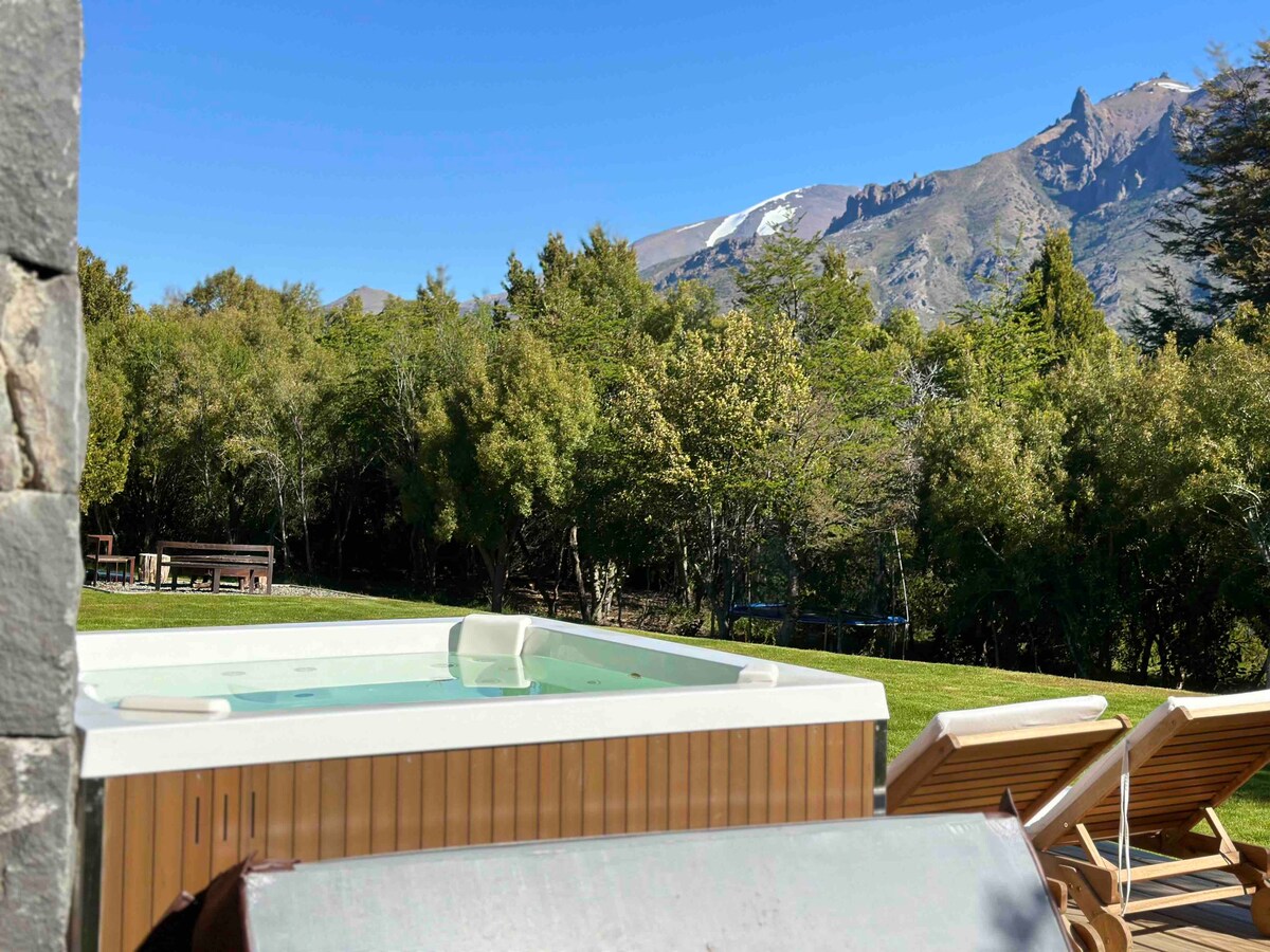 An outdoor jacuzzi is positioned in a serene garden, surrounded by lush greenery. In the background, a mountain range features snow-capped peaks, offering a striking natural backdrop under a clear blue sky. Sun loungers are placed nearby for relaxation.