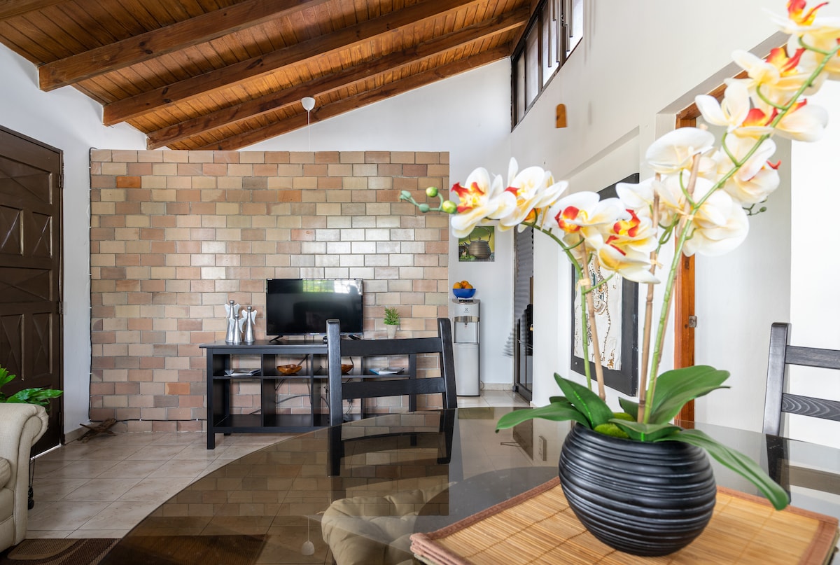 A bright living area features a decorative wall with a warm brick design, complemented by a wooden ceiling. A television is positioned on a simple black console, while a round dining table is adorned with a vase of elegant flowers, adding a touch of nature.