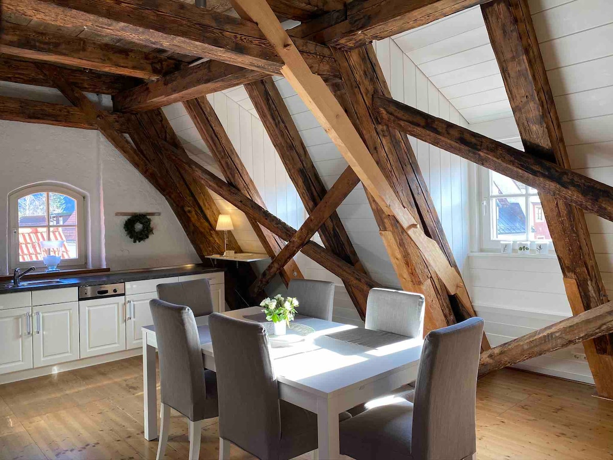 A bright dining area is highlighted by exposed wooden beams and large windows. A white table is surrounded by four grey upholstered chairs. The kitchen area features white cabinetry and is partially visible in the background.