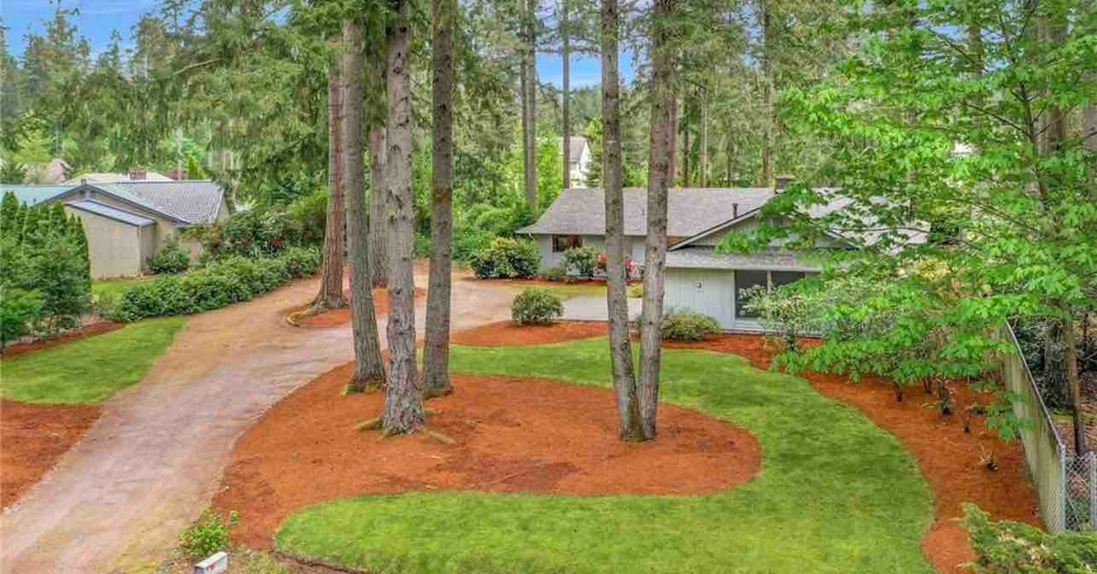 The landscaped exterior features a winding pathway among lush green grass and trees. Soft brown mulch outlines garden beds filled with vibrant foliage, contributing to a serene outdoor environment. A peaceful residential setting is evident, with neighboring homes nestled among the trees.