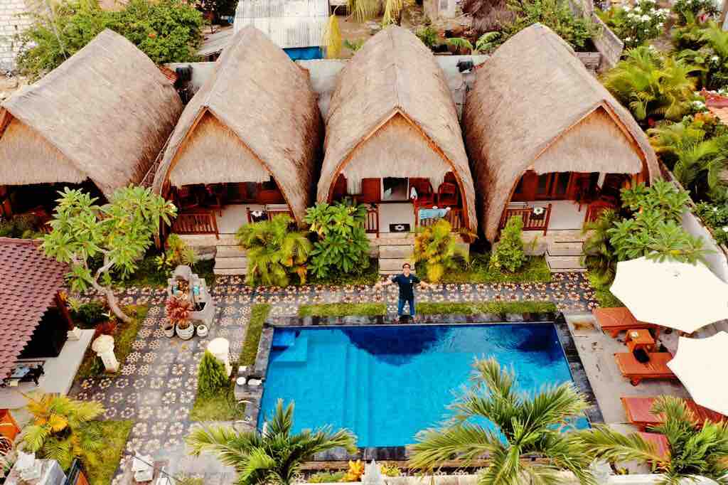 Aerial view of a vibrant outdoor area featuring three thatched-roof bungalows arranged around a rectangular pool. Lush greenery surrounds the pool, and a patterned stone pathway leads to seating areas with umbrellas. Sunlight reflects off the clear blue water, inviting relaxation.