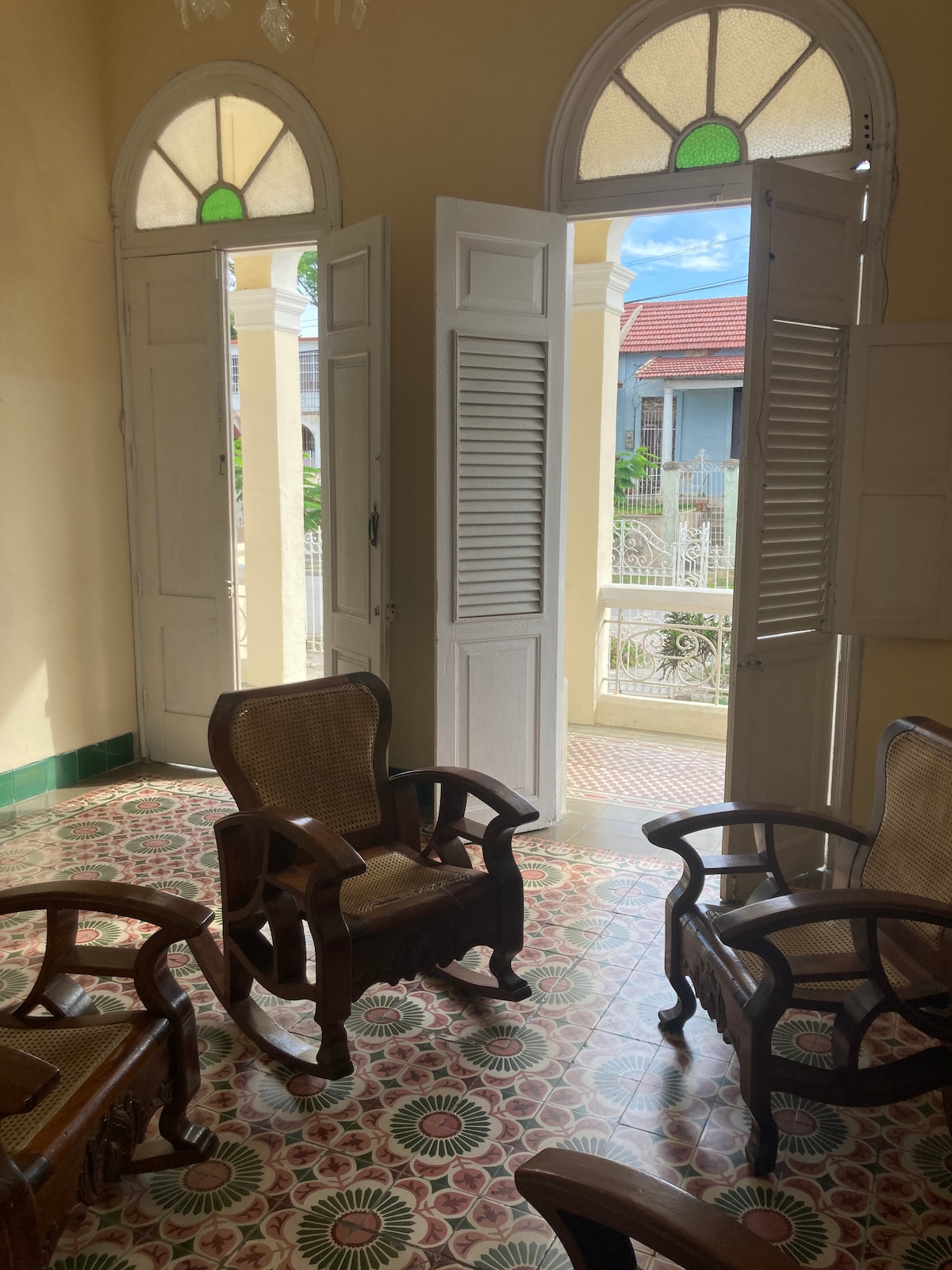 Two wooden chairs with woven seats are positioned on a patterned tiled floor. Open French doors with louvered shutters lead to an outdoor balcony, allowing natural light to fill the room. The walls are painted in a light color, enhancing the airy feel.