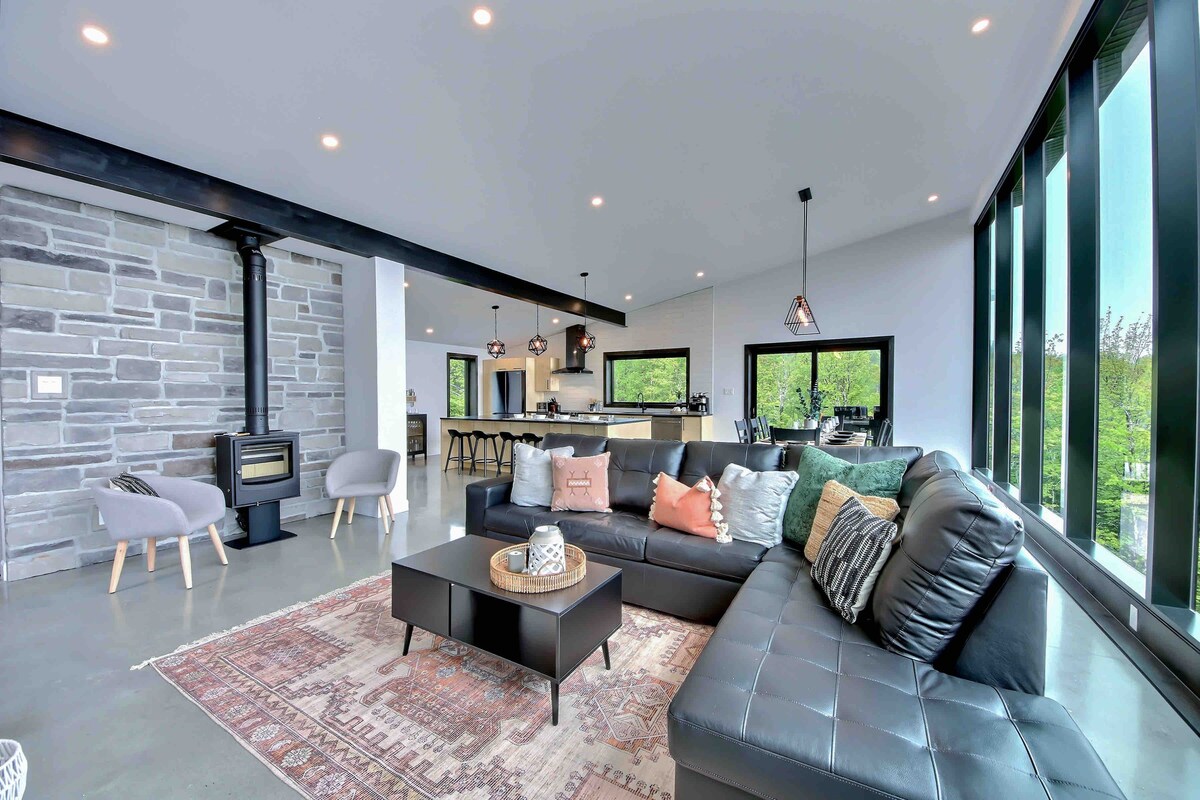 A spacious living area features a modern sectional sofa and a coffee table on a decorative rug. A stone accent wall complements large windows, providing ample natural light. A dining area with high stools and a view of greenery is visible in the background.