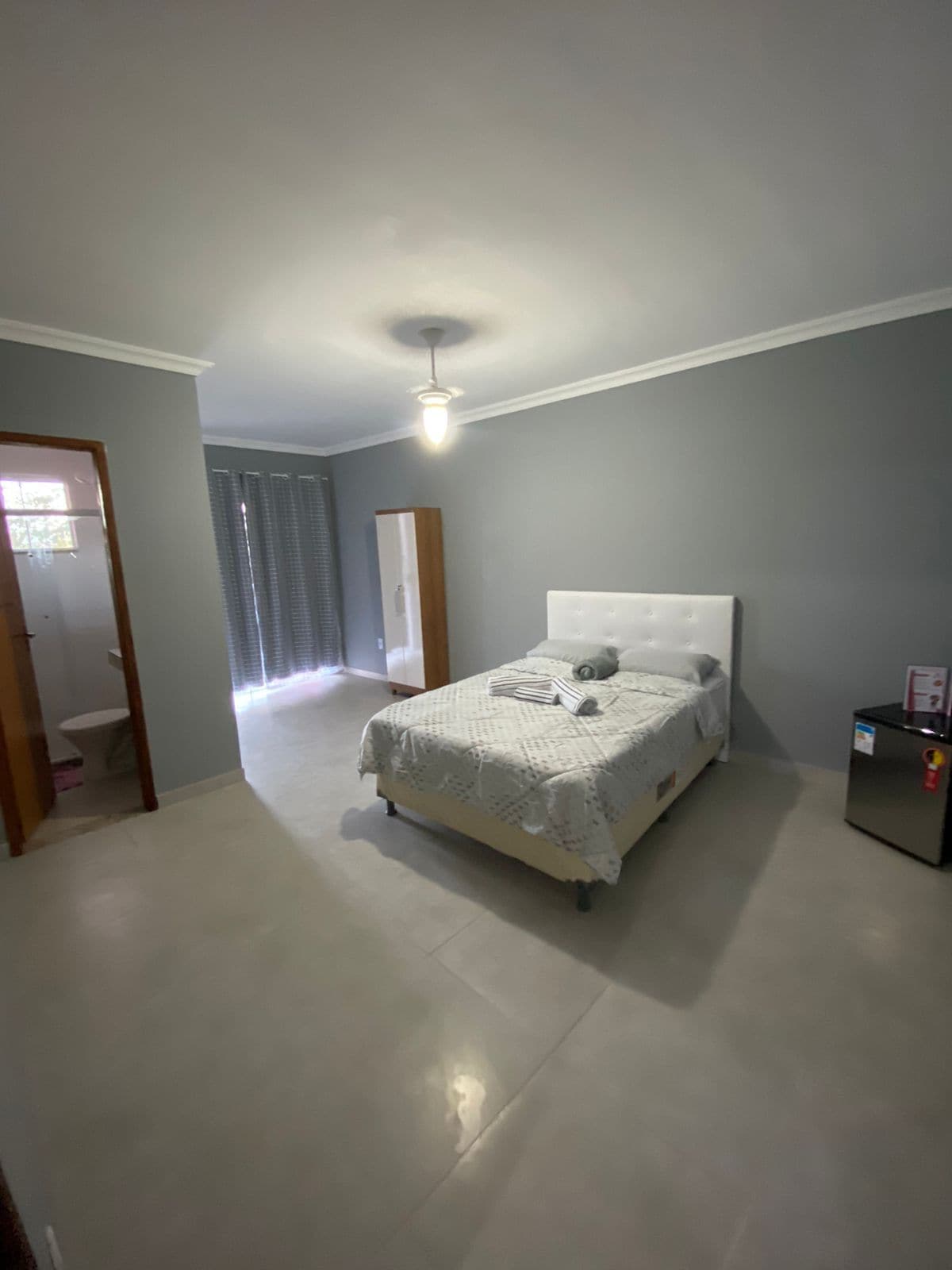 A cozy bedroom is furnished with a spacious bed covered in light bedding. A window with blinds allows natural light to enter, while a small refrigerator and wardrobe are positioned nearby. The room features a simple, neutral color palette with polished flooring.