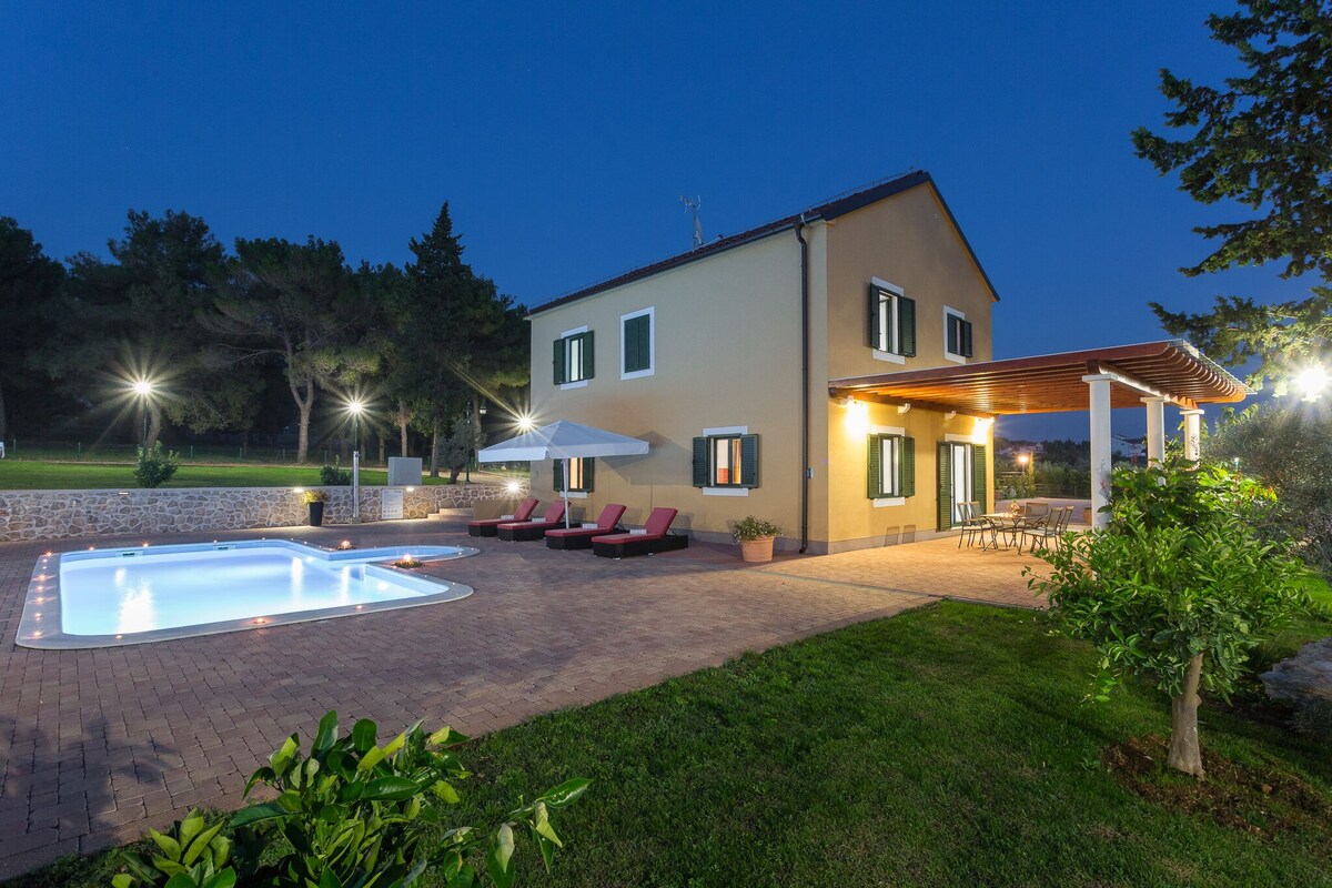 The villa is illuminated at night, showcasing an outdoor area featuring a pool surrounded by sun loungers. A spacious terrace is visible with dining options, framed by lush greenery and trees, creating a serene environment. Soft lighting enhances the inviting atmosphere of the property.