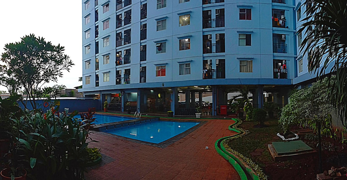 A serene outdoor area features a swimming pool surrounded by lush greenery and landscaped pathways. The multi-story building stands in the background, with multiple balconies visible. Soft evening light casts a gentle glow over the scene, enhancing the calm atmosphere of the space.