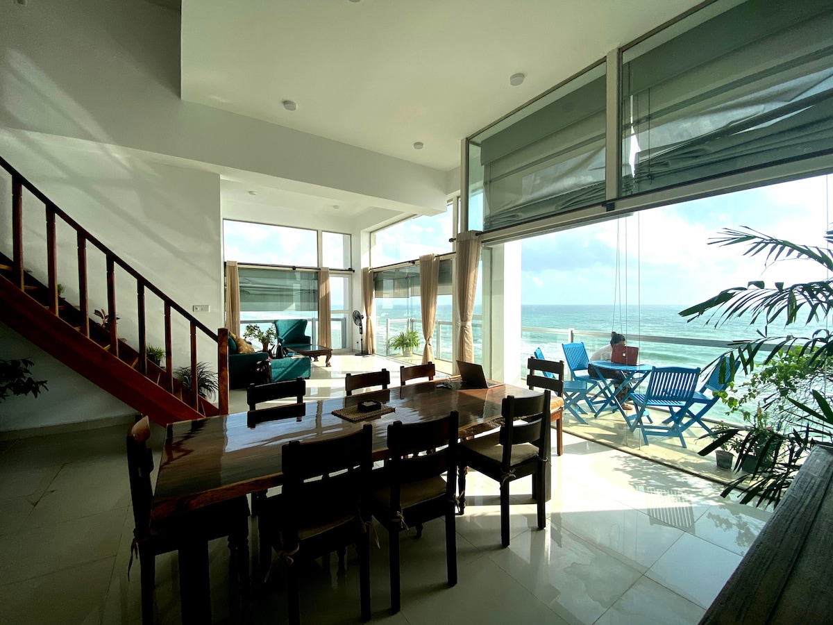 A spacious living and dining area is visible, featuring a long wooden table surrounded by chairs. Large glass doors reveal a sea view and a seating area on the terrace. Natural light fills the space, complemented by minimalist decor and plants.