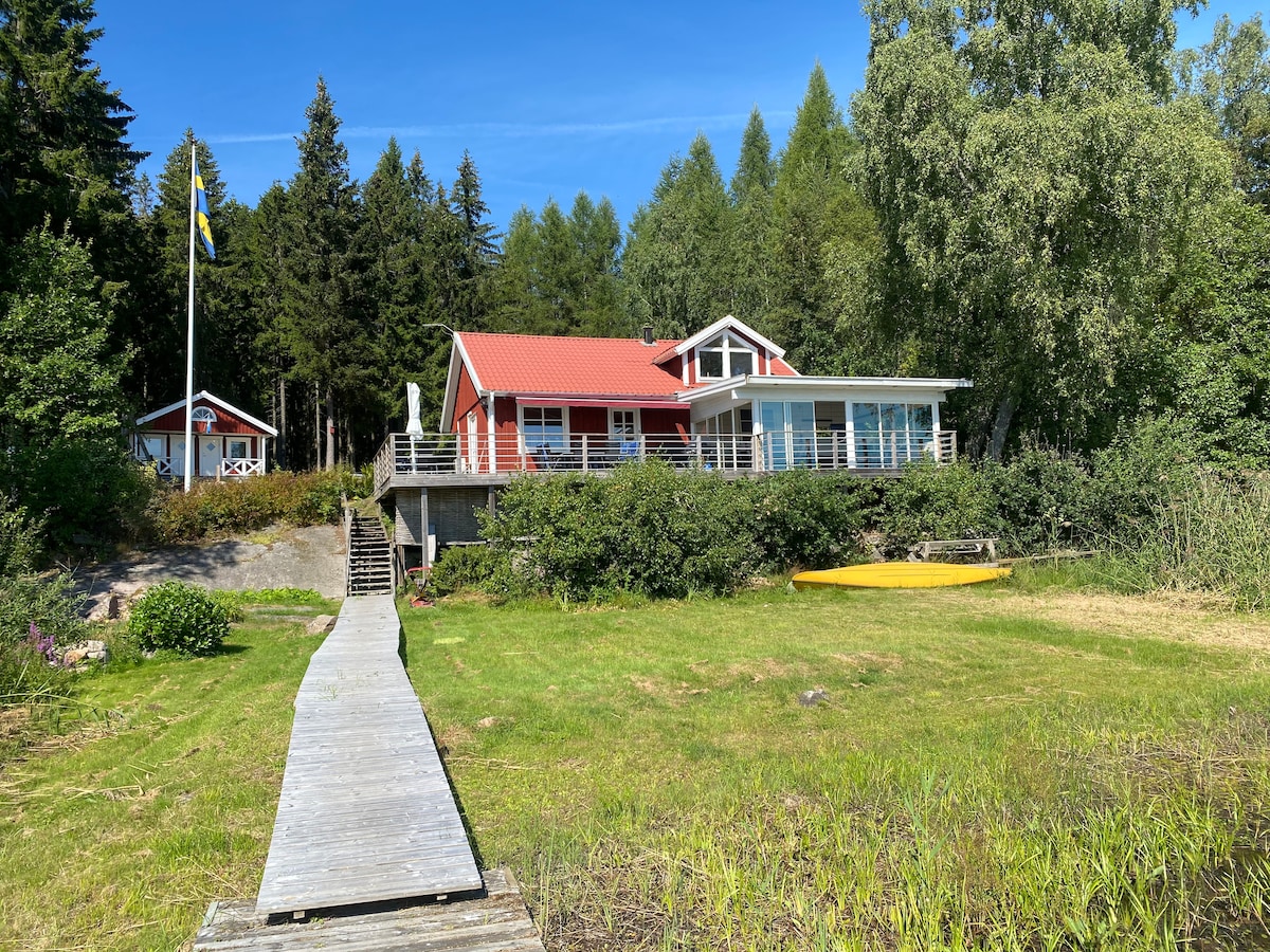A lakeside cabin is set against a backdrop of lush greenery and trees, featuring a red exterior and a welcoming porch. A wooden pathway leads to the water's edge, where a yellow kayak is visible, providing an invitation to outdoor activities.