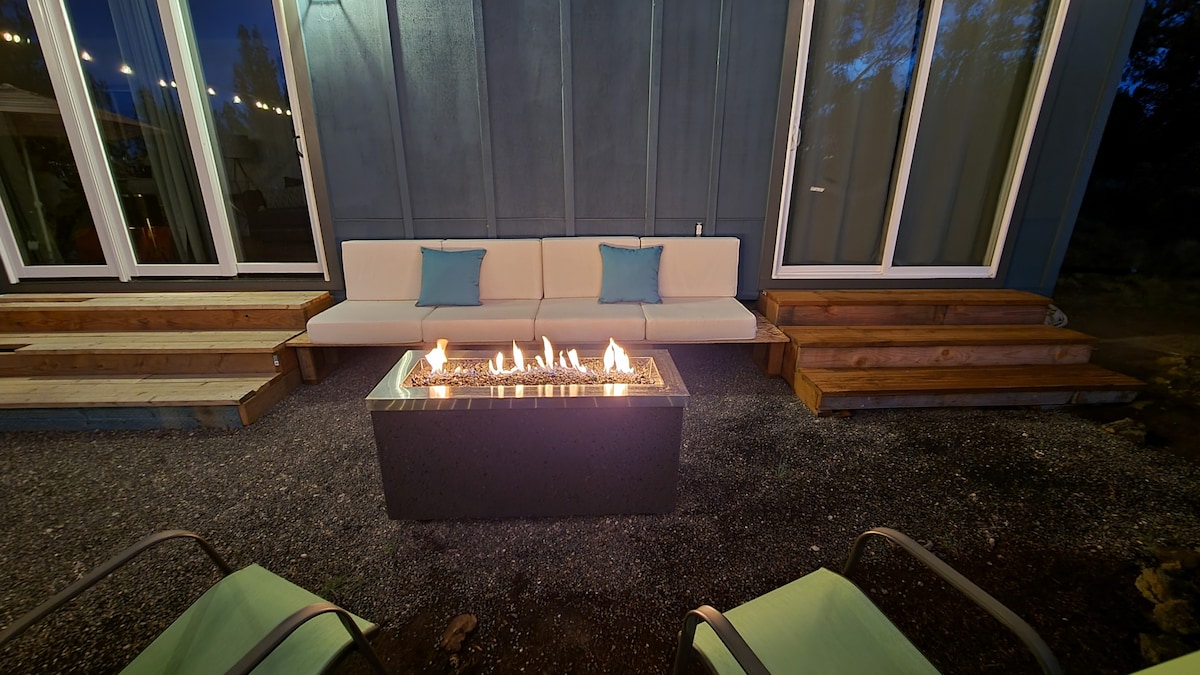 A cozy outdoor seating area features a modern fire pit surrounded by comfortable couches with decorative cushions. Soft lighting enhances the inviting atmosphere, and surrounding greenery complements the space, making it suitable for relaxation and social gatherings.