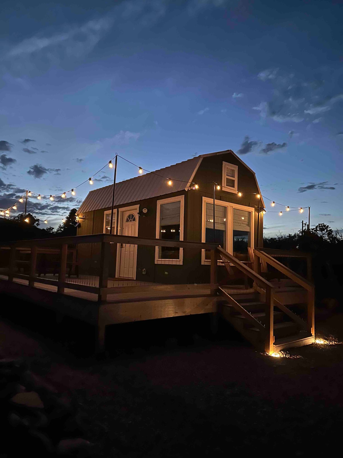 The tiny home is illuminated by string lights, creating a warm ambiance against the twilight sky. A spacious deck features wooden railings and stairs leading up to the entrance, enhancing the inviting atmosphere of the retreat.