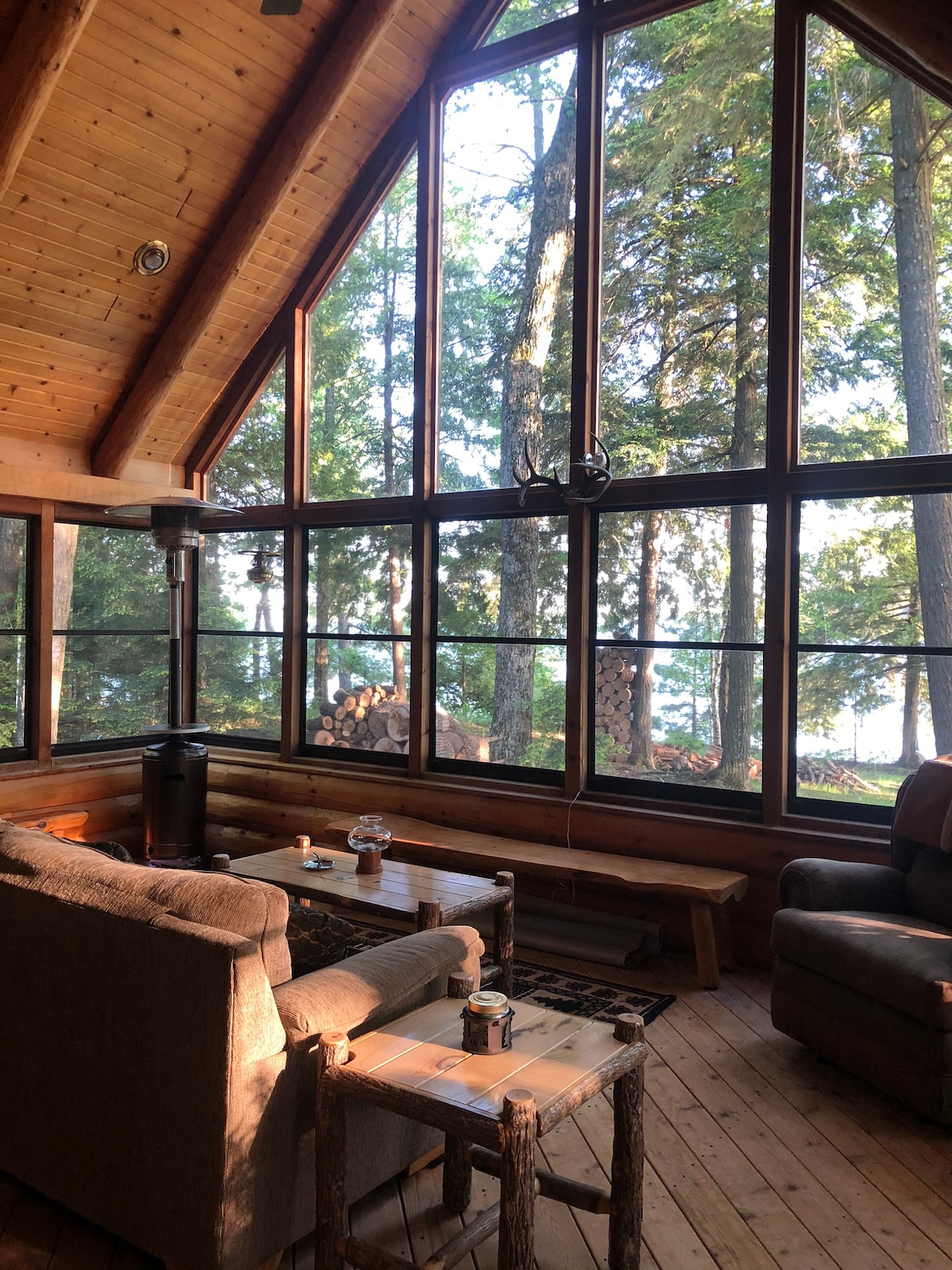 A bright living space features large windows showcasing views of tall trees and the water beyond. A cozy seating area includes a sofa and armchair arranged around a wooden coffee table. Natural wood finishes create a warm ambiance, complemented by a small decorative lamp on the table.