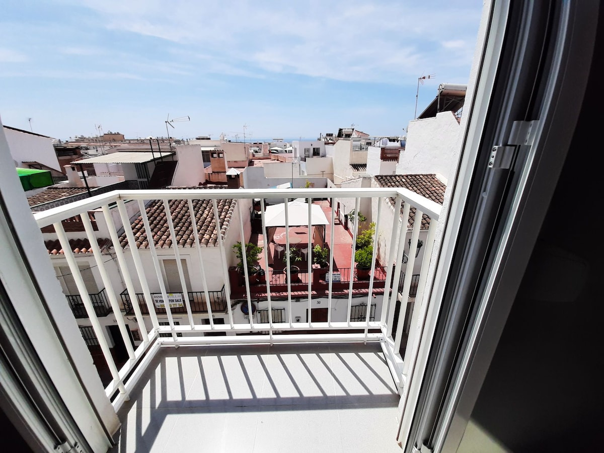 A juliet balcony is visible, offering a partial view of the sea and surrounding rooftops. The balcony features a white railing, with sunlight casting shadows on the floor from the open doors. A bright blue sky adds to the welcoming atmosphere.