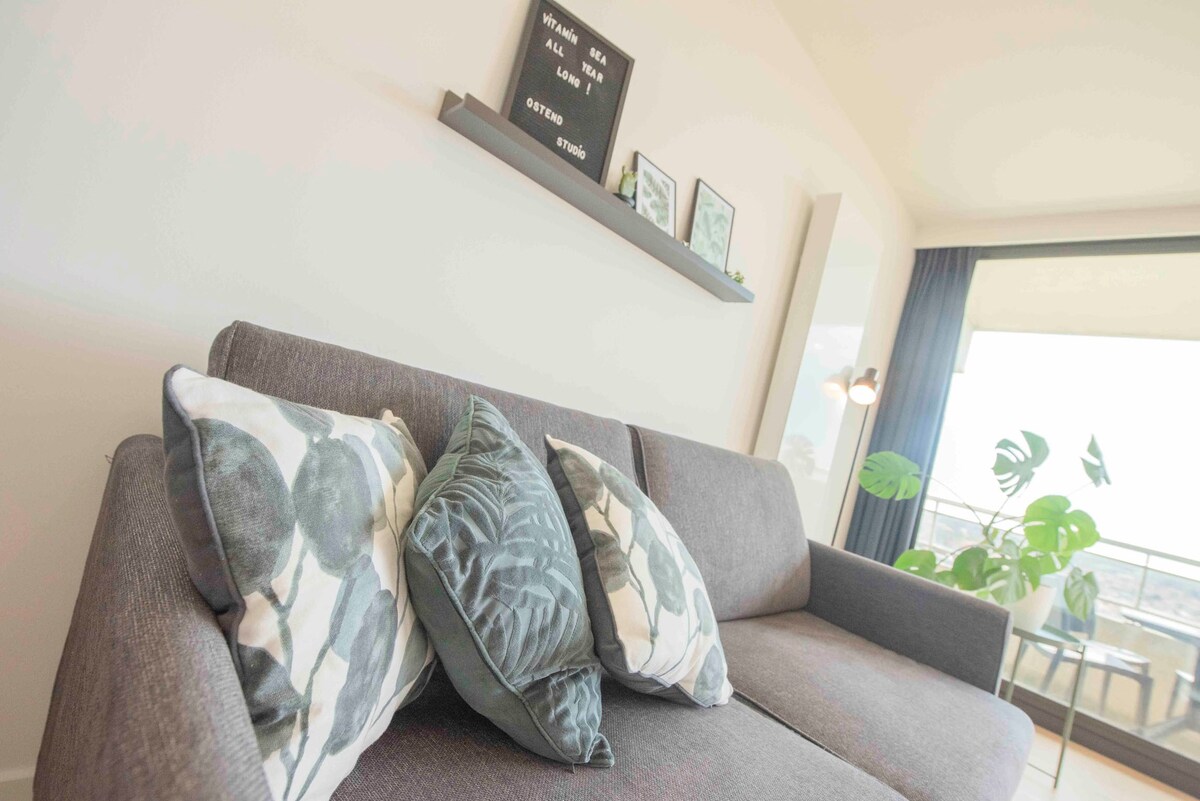 A modern seating area is highlighted with a gray sofa adorned with patterned cushions in green and blue hues. Above the sofa, a minimalist shelf displays decorative items. Natural light enters through large windows, enhancing the calm ambiance.