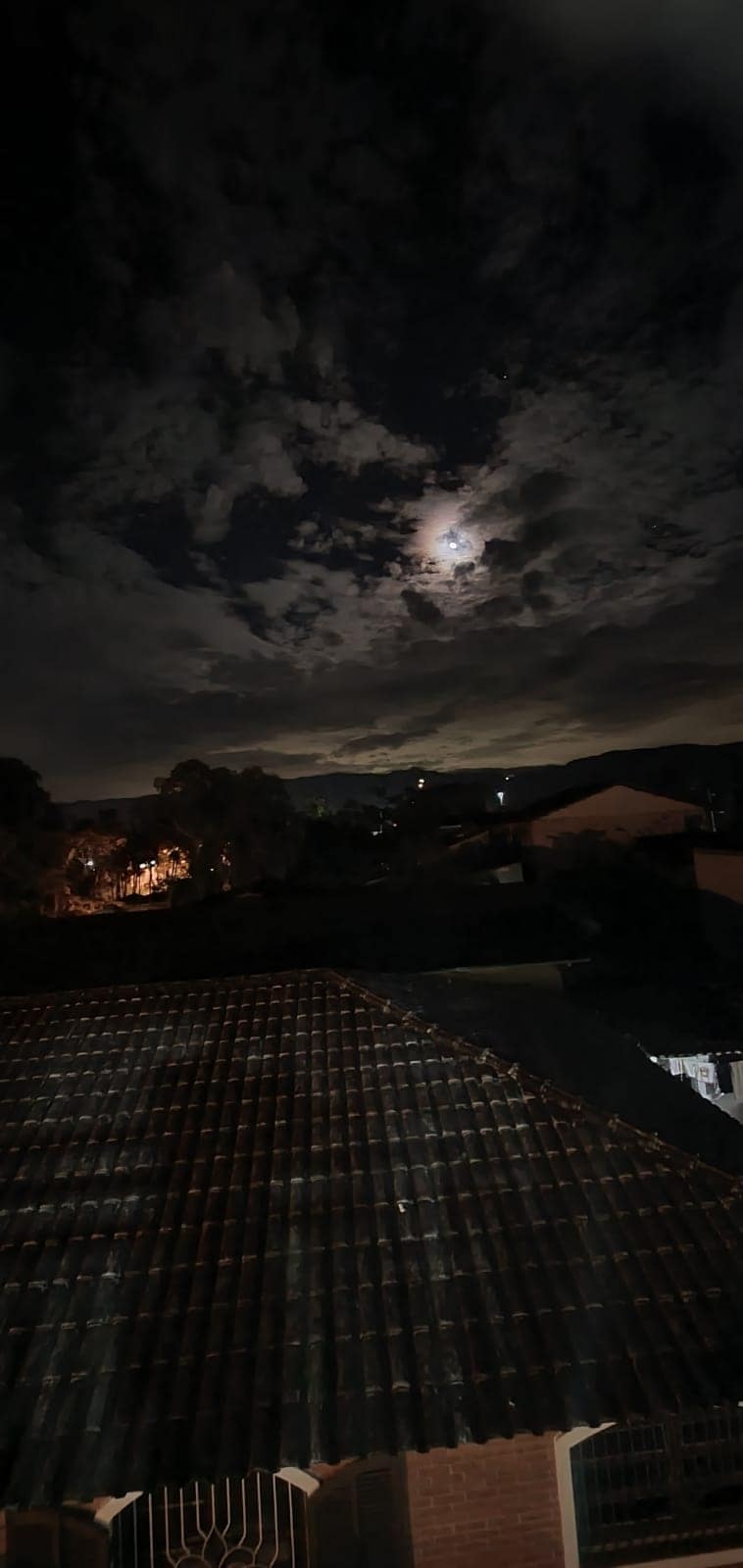 A nighttime view captures a moonlit sky filled with scattered clouds, creating soft contrasts. The silhouette of nearby houses and trees is visible, with warm lights emanating from structures below, contributing to the serene ambiance of the landscape.
