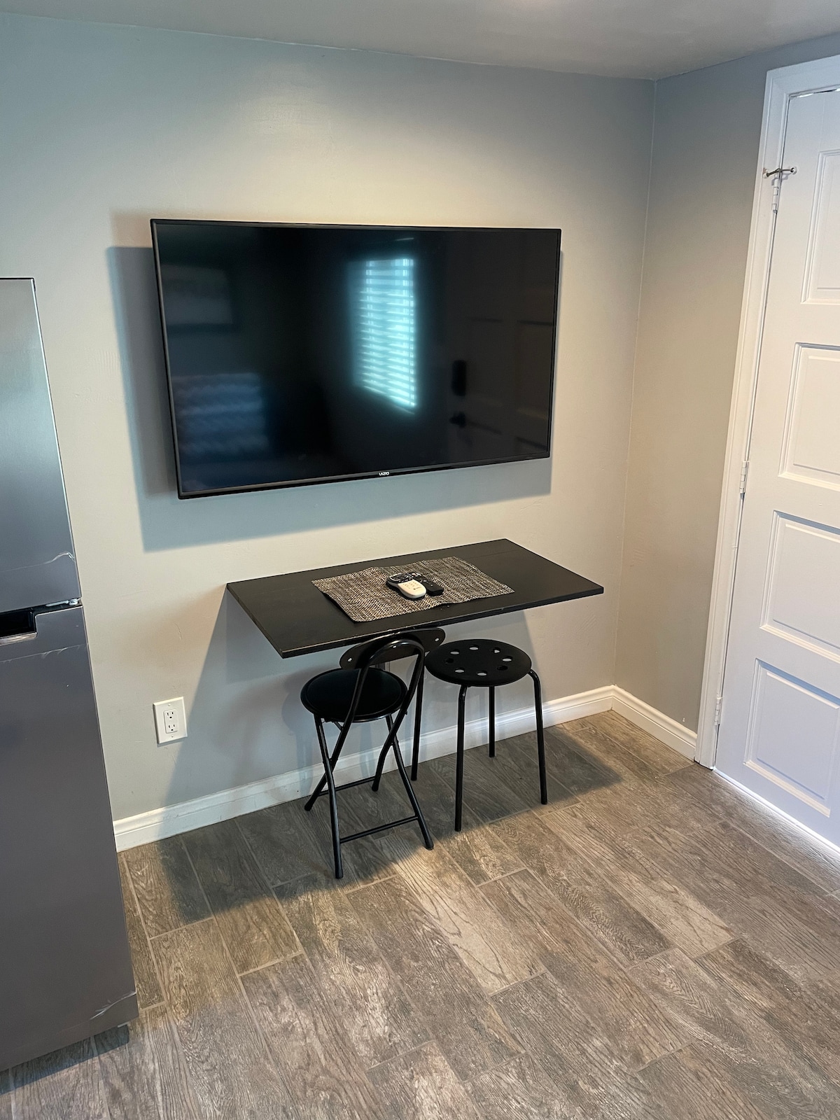 A modern flat-screen TV is mounted on the light gray wall, providing entertainment options. Below, a small black table is paired with two sleek, black chairs. The flooring is tiled in a light gray tone, enhancing the clean design of the space.