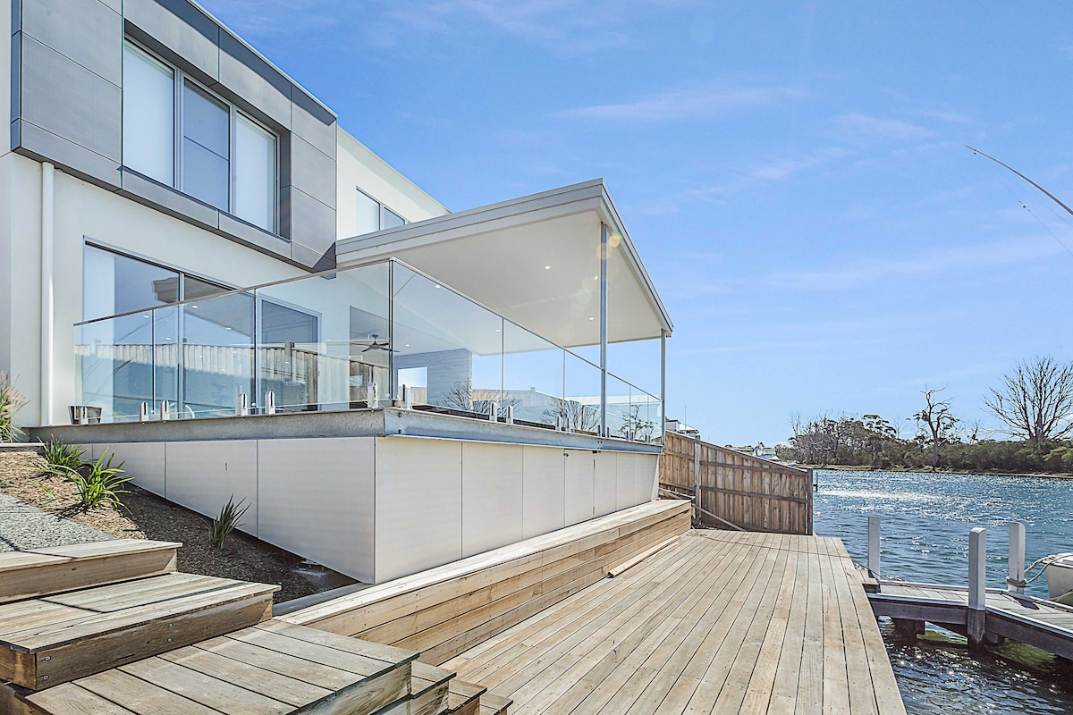 The waterfront deck extends from the contemporary home, featuring glass balustrades and overlooking the canal. Sunlight reflects off the water, highlighting the wooden walkway that connects the deck to the jetty, enhancing the connection to the surrounding natural beauty.