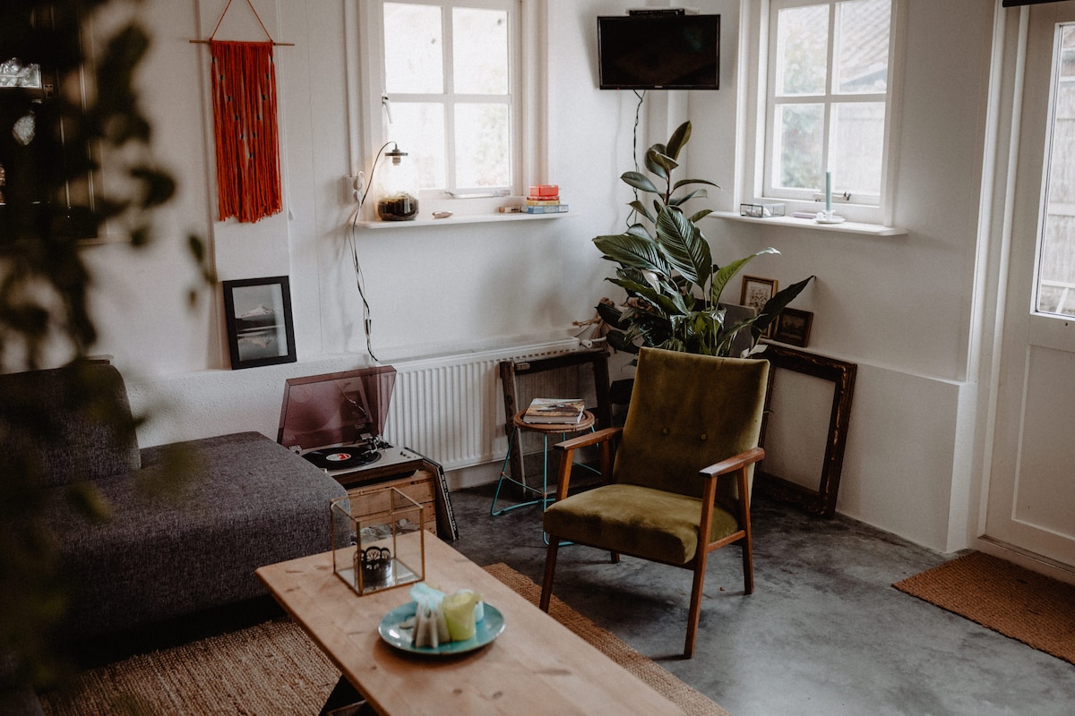 A relaxed living space is presented with a green accent chair and a gray sofa. A wooden coffee table is placed in front of the sofa, while a record player and framed artwork add character. Natural light enters through the windows, enhancing the cozy environment.