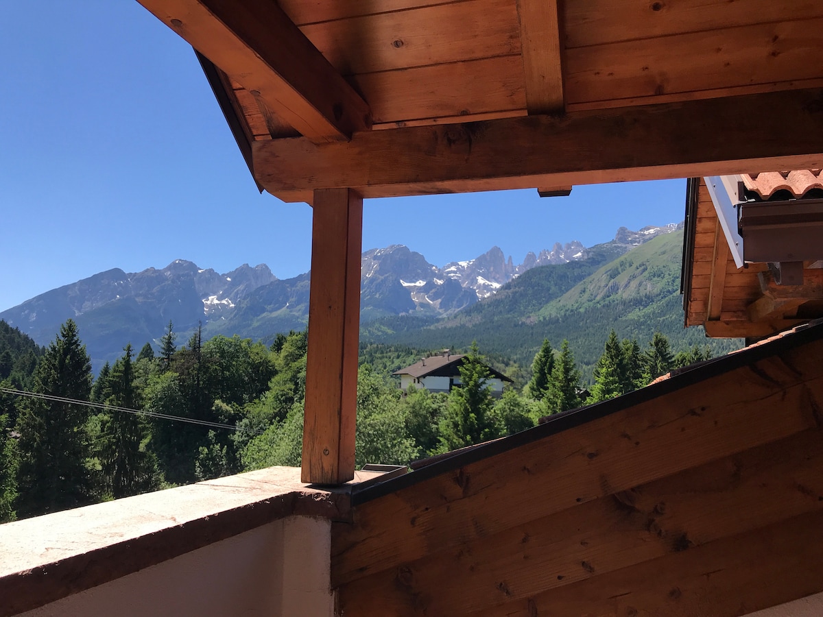 A view from the balcony showcases the surrounding alpine landscape framed by wooden beams. The majestic Dolomites rise in the background, while lush greenery is visible in the foreground, offering a serene mountainous atmosphere.