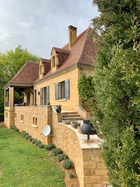The stone house is framed by lush greenery, showcasing a sloped roof and intricate architectural details. A terraced area leads to a spacious porch, where potted plants and a traditional cooking pot are placed on the stone steps.