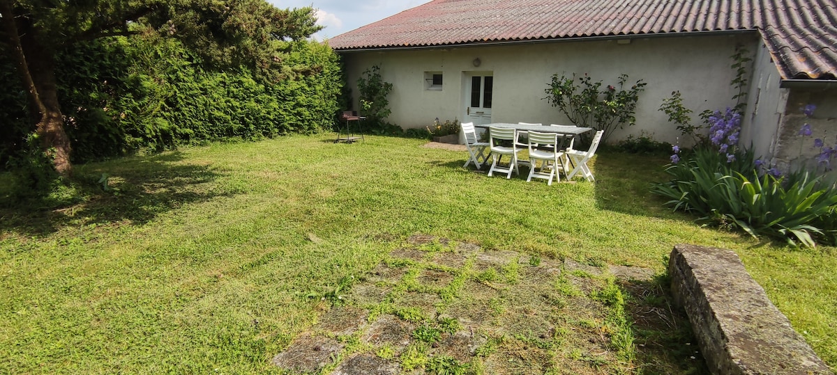 A spacious garden area is depicted, featuring a well-maintained lawn and a patio table set with several chairs. The garden is bordered by lush greenery and flowering plants. A barbecue grill is positioned nearby, alongside the exterior wall of the house.