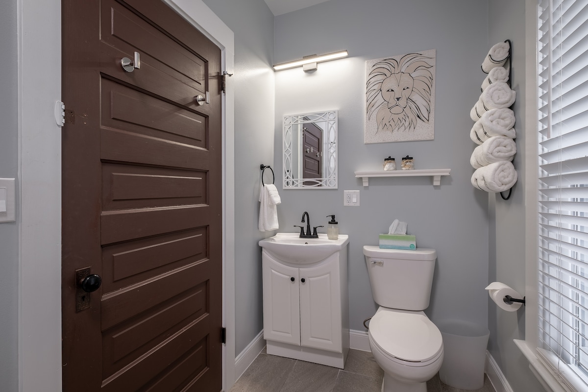 The newly remodeled bathroom includes a sink with a large mirror above it, a toilet, and a towel rack. Fresh towels are neatly arranged on the rack. A decorative wall art piece featuring a lion adds character to the light grey walls.