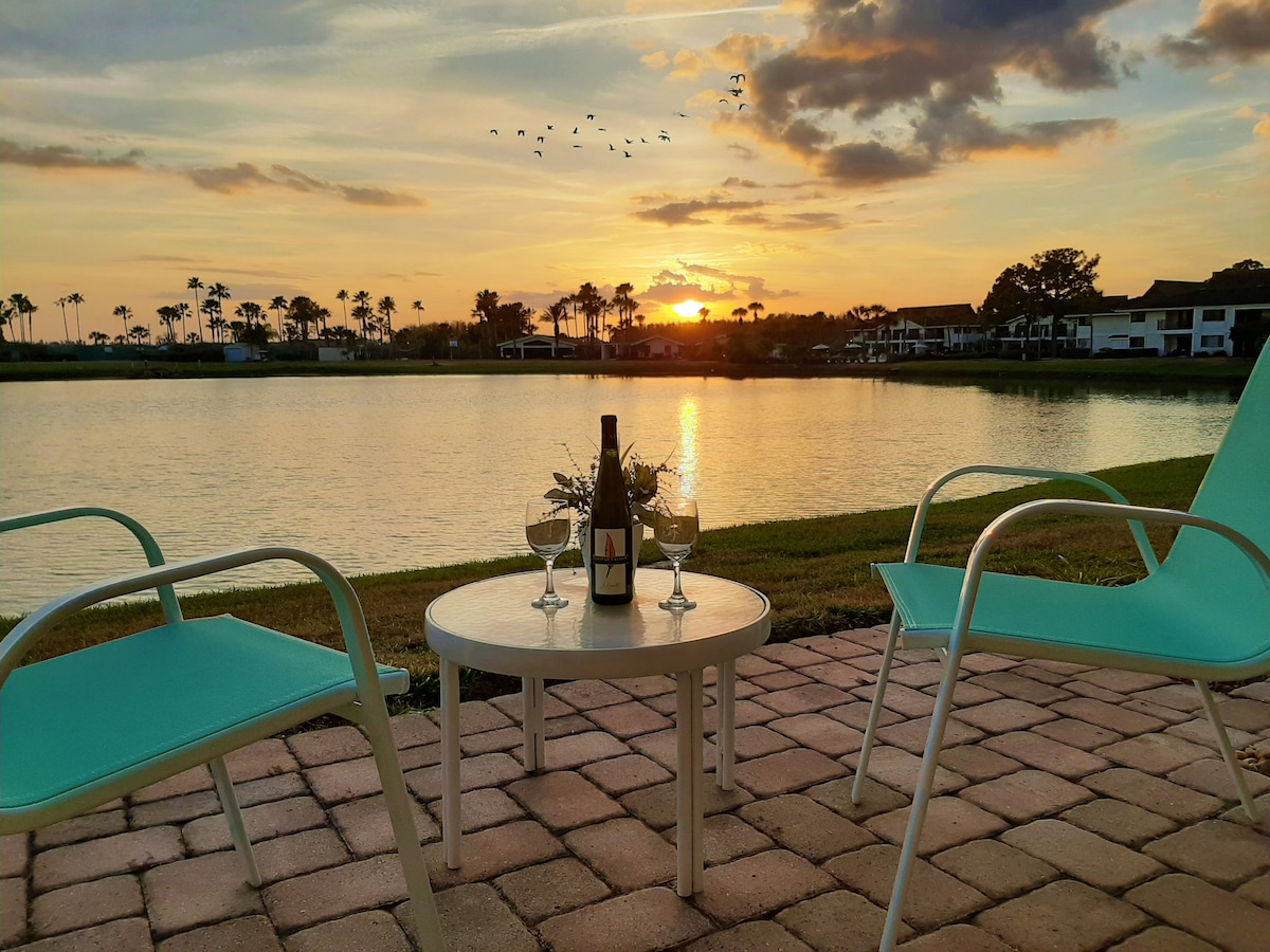 A serene terrace scene showcases a small table set with a wine bottle and two glasses, framed by two aqua-colored chairs. The calm lake reflects a vibrant sunset, while palm trees outline the horizon in the distance. A flock of birds is seen flying across the sky.